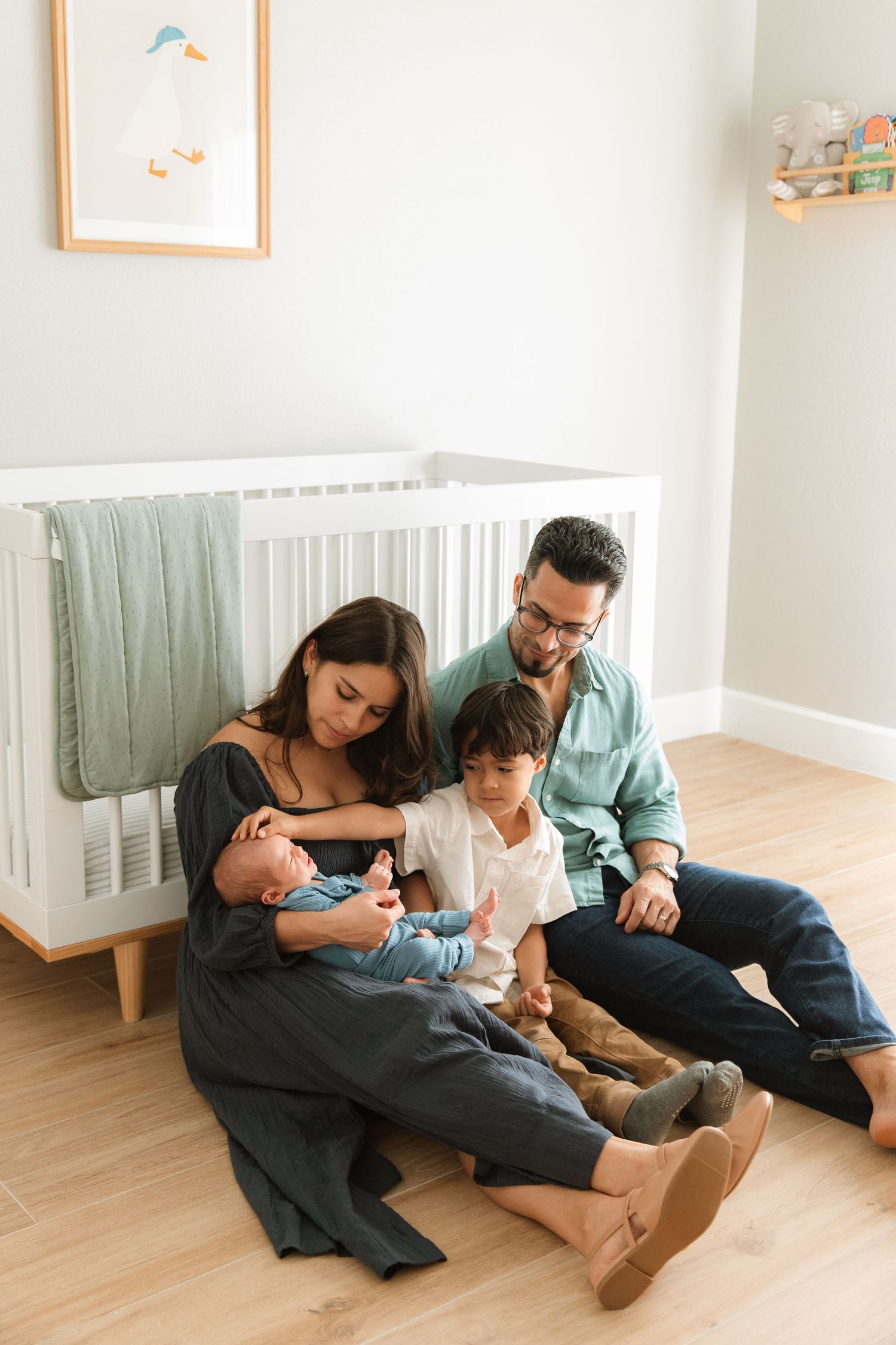 Family of four sitting on nursery floor near white crib with newborn baby.