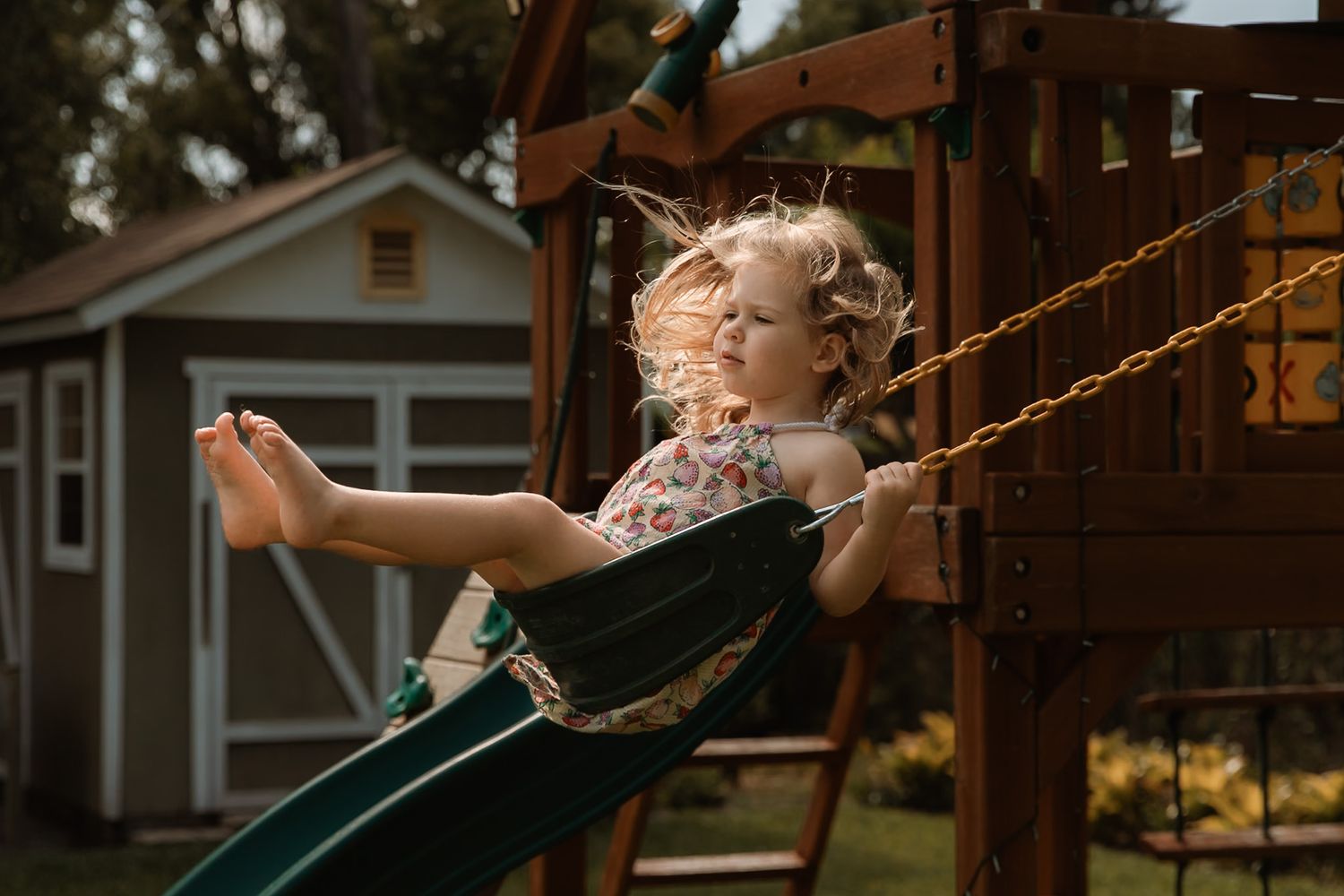A young blonde girl joyfully rides a swing on a backyard playset during golden hour.