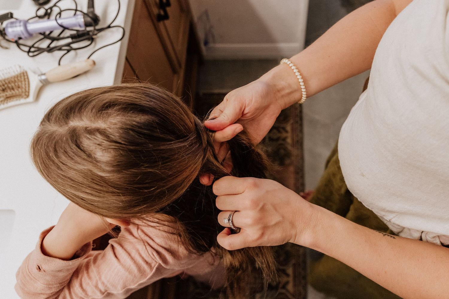 Mother braiding young daughters long brown hair in a bright bathroom setting.
