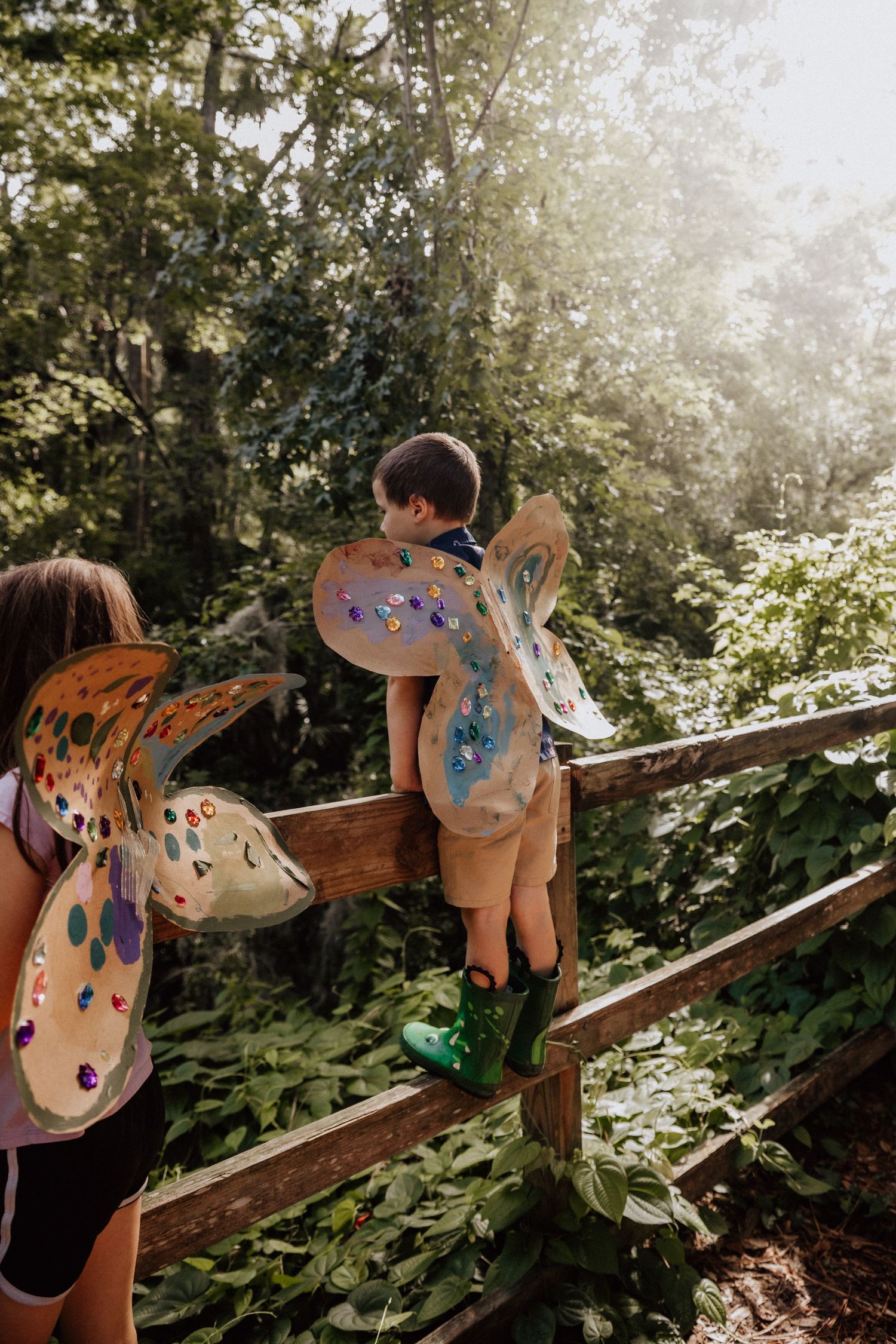 Two children wearing colorful handmade butterfly wings explore a wooden fence in a sunlit forest.