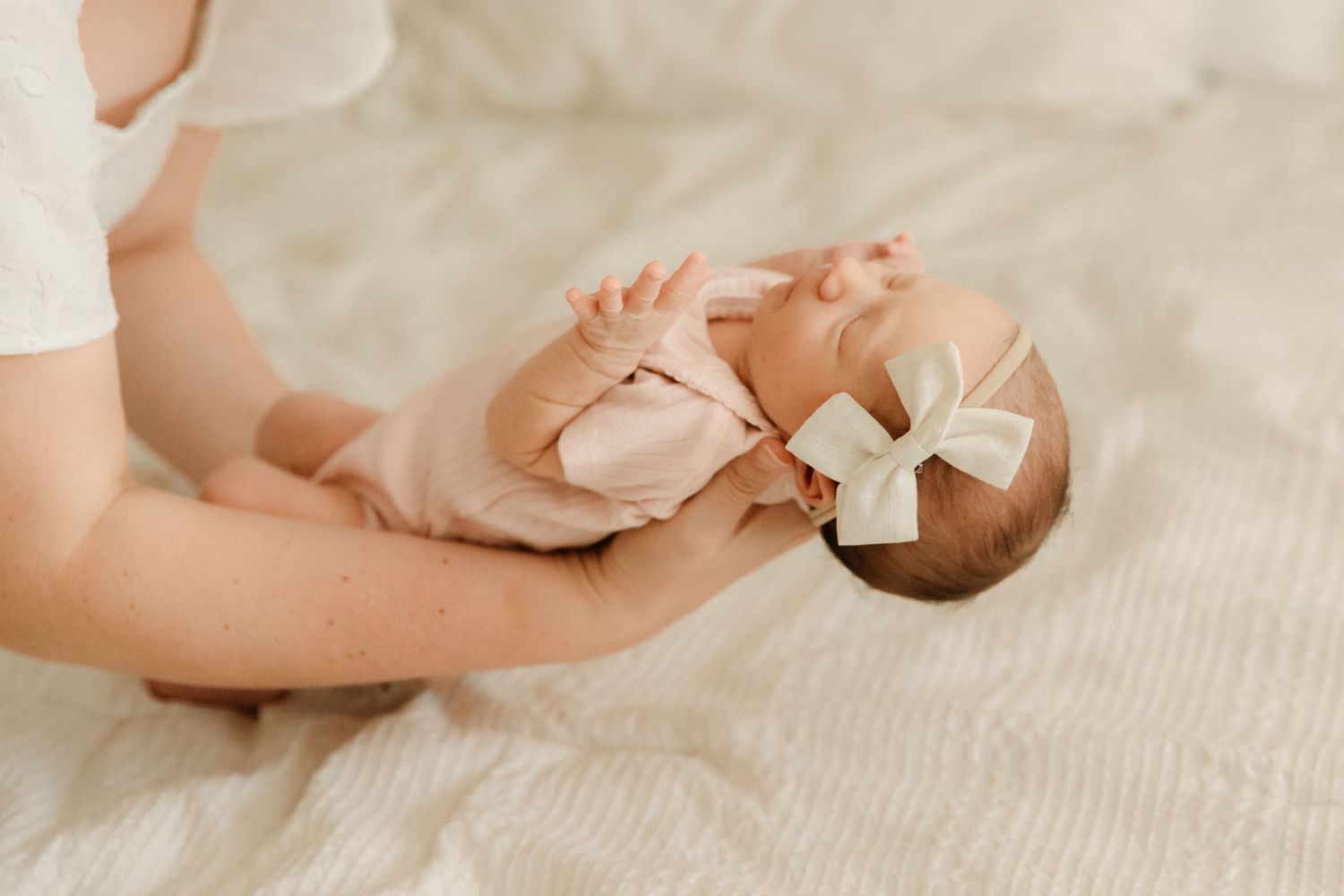 A newborn baby in a pink onesie and white bow headband rests in a parent's hands on a soft white blanket.