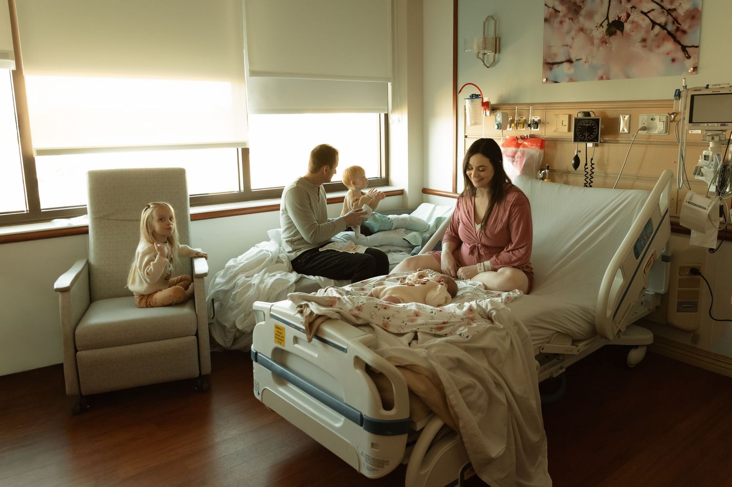 Family in hospital room with newborn baby, mother resting in bed while father and older children sit nearby.