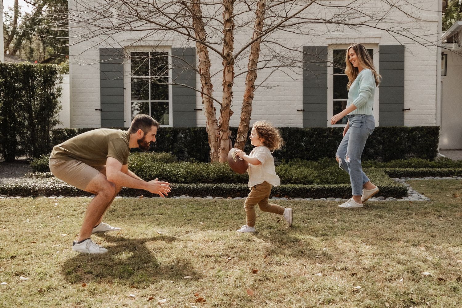 Young family playing in yard, father crouching as toddler runs toward him with mother nearby.