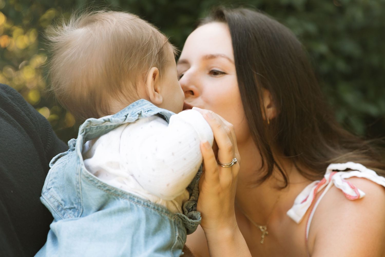A mother playfully kisses her baby dressed in denim outdoors in natural sunlight.
