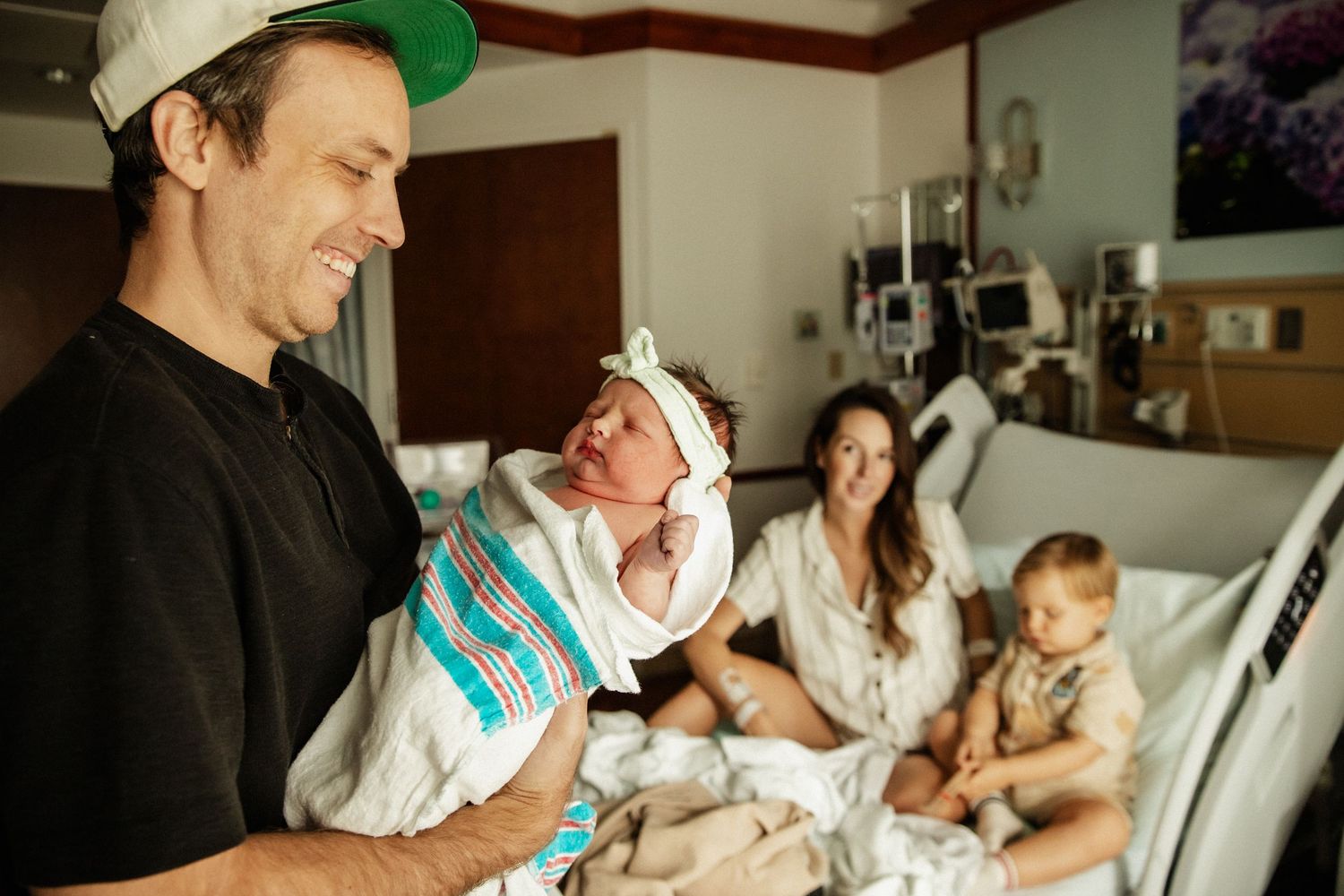Proud father holds swaddled newborn in hospital room as mother and toddler sit on bed.