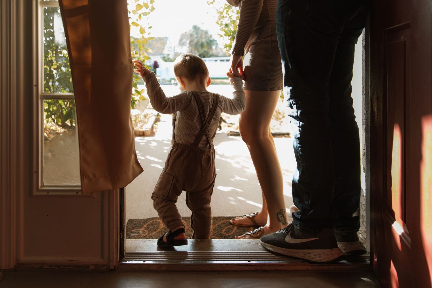 Silhouette of toddler in suspenders standing at open front door with parent beside them.