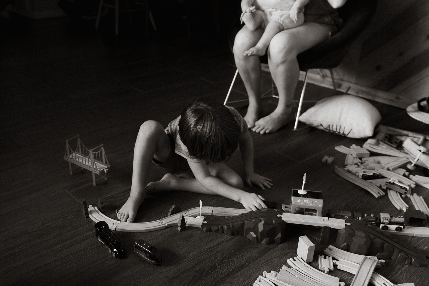 Black and white photo of young child playing with wooden train set on floor.