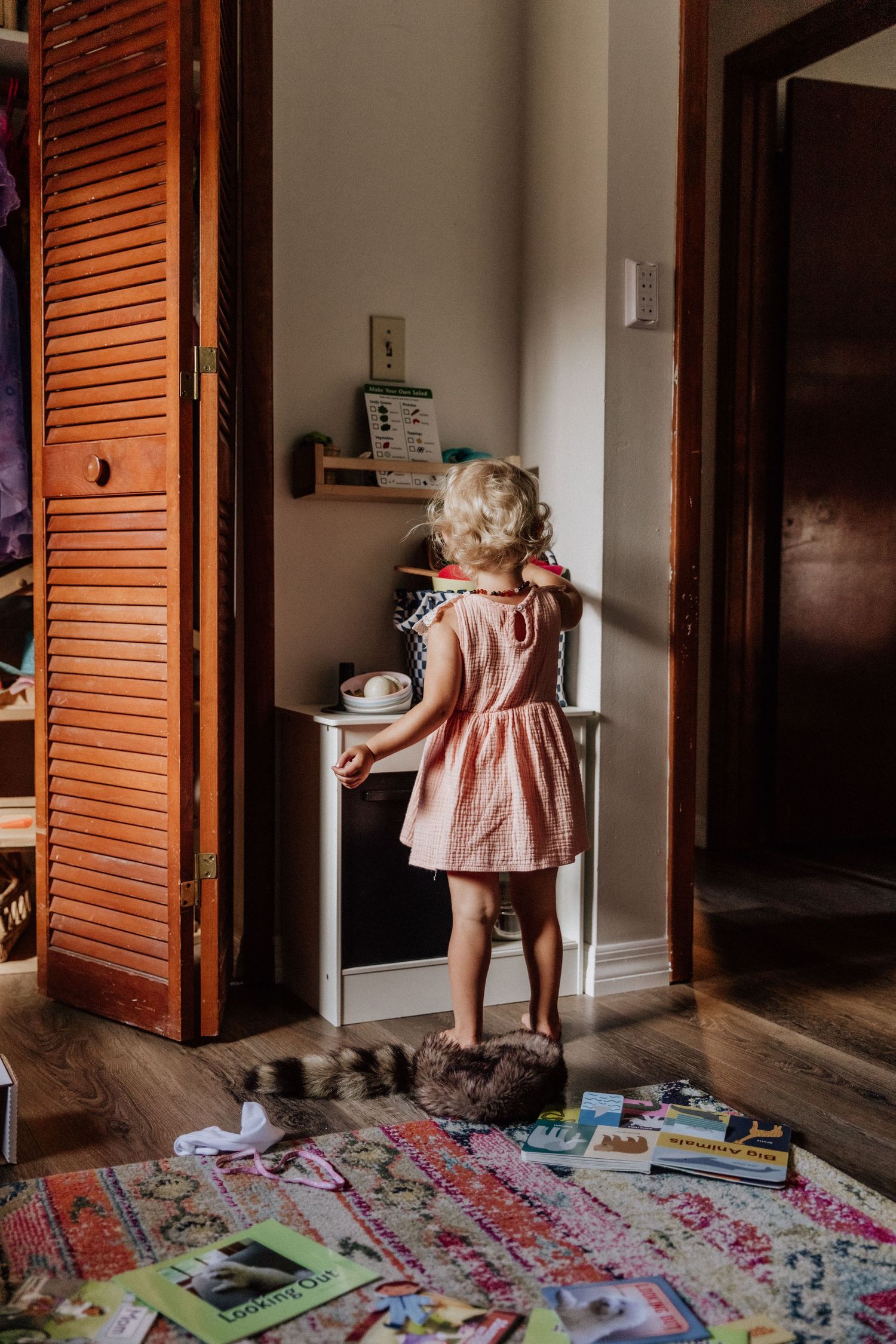 Young child in pink dress reaching for light switch with toys scattered on patterned rug below.
