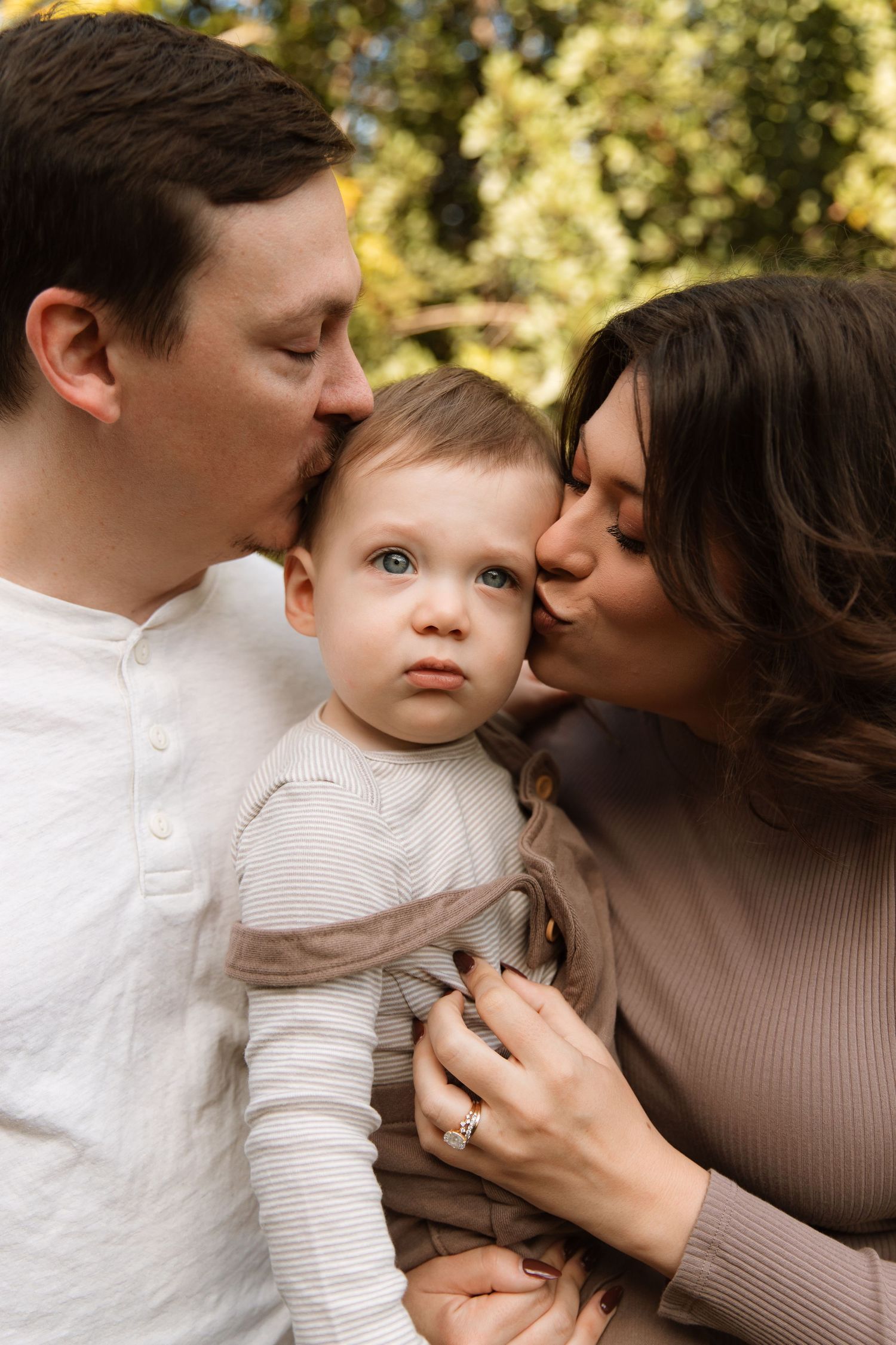 Parents kissing their baby on both cheeks simultaneously outdoors in autumn setting.