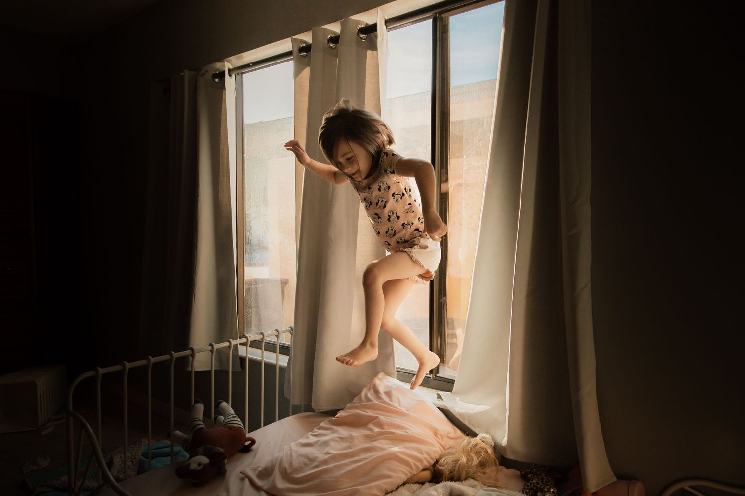Young girl jumping energetically on a bed near a bright window in a warmly lit bedroom.