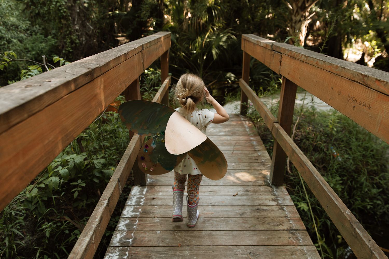 A young child wearing fairy wings walks across a wooden bridge surrounded by lush greenery.