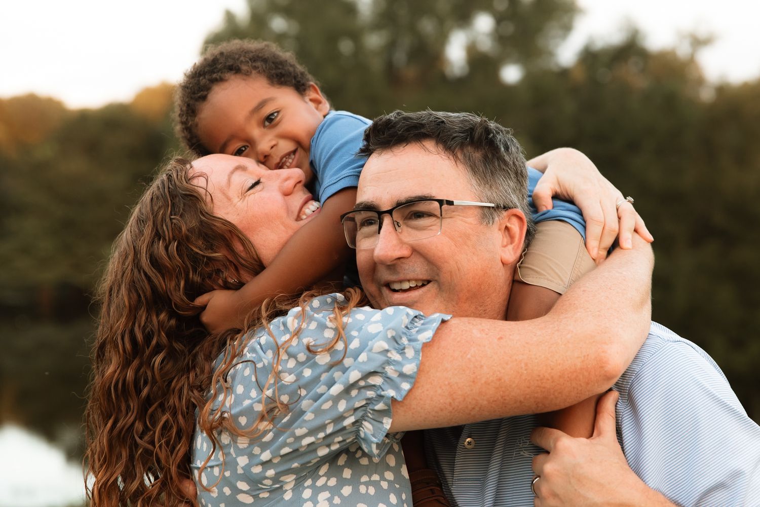Joyful family moment as a child hugs both smiling parents outdoors near a peaceful lake setting.