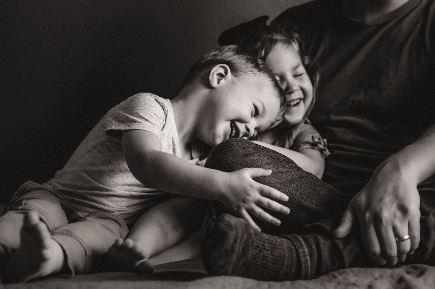 Black and white photo of two laughing children cuddling with parent on bed.