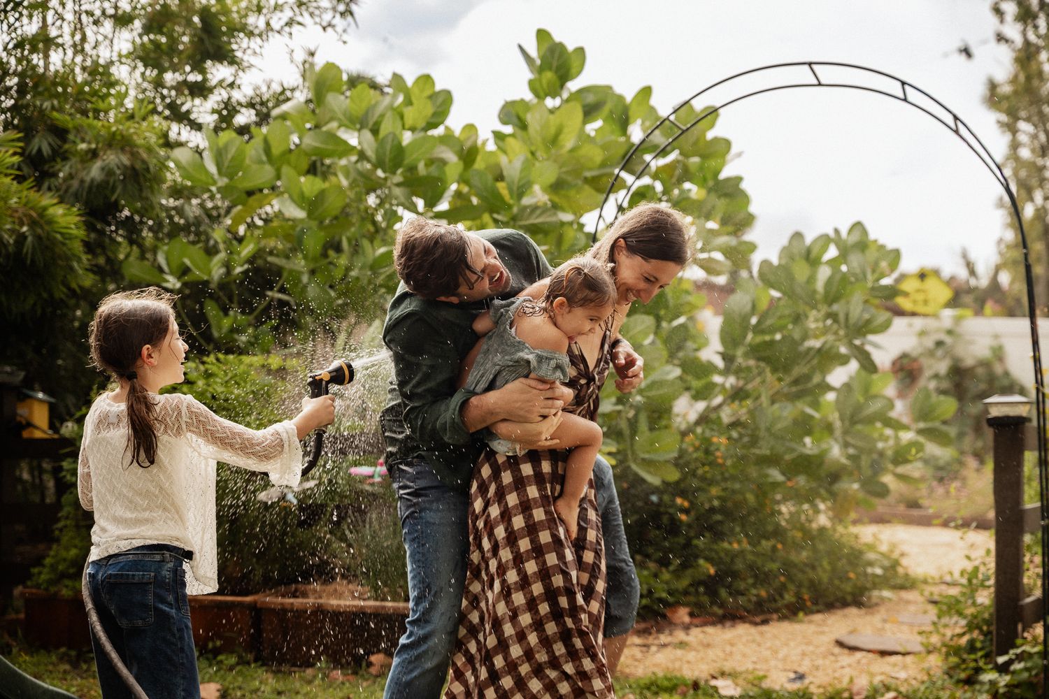 A joyful family plays together in a lush garden as a child sprays water from a hose.