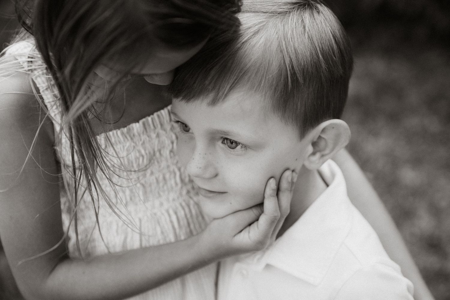 Black and white photo of an older girl tenderly cradling a young boys face in her hands.