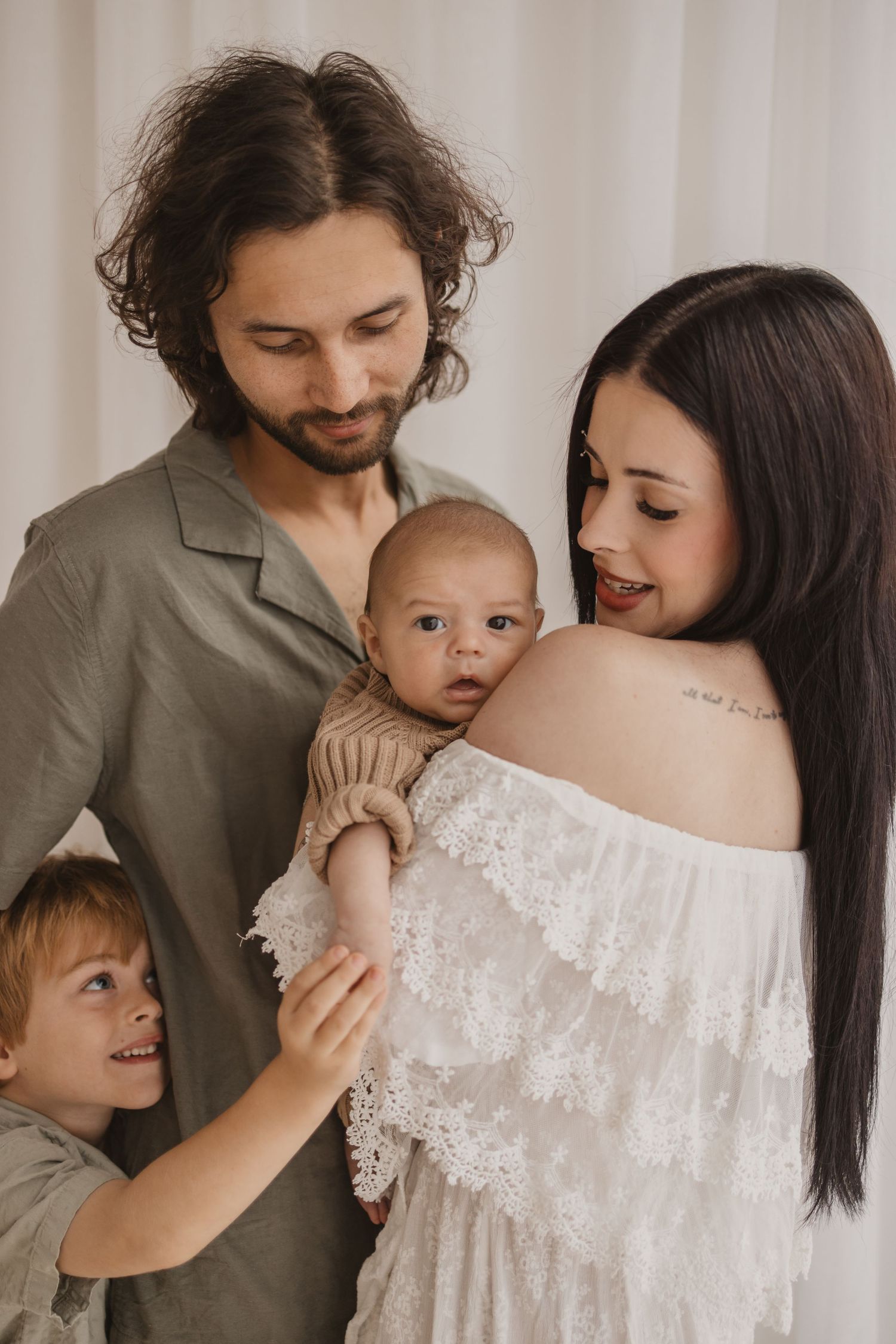 A young family with a bearded father, dark-haired mother, toddler son, and newborn baby posing together indoors.