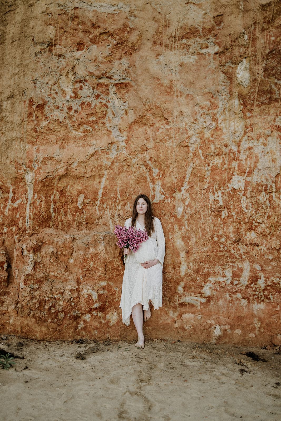 Pregnant woman in white dress holding pink flowers against rustic red clay wall outdoors.