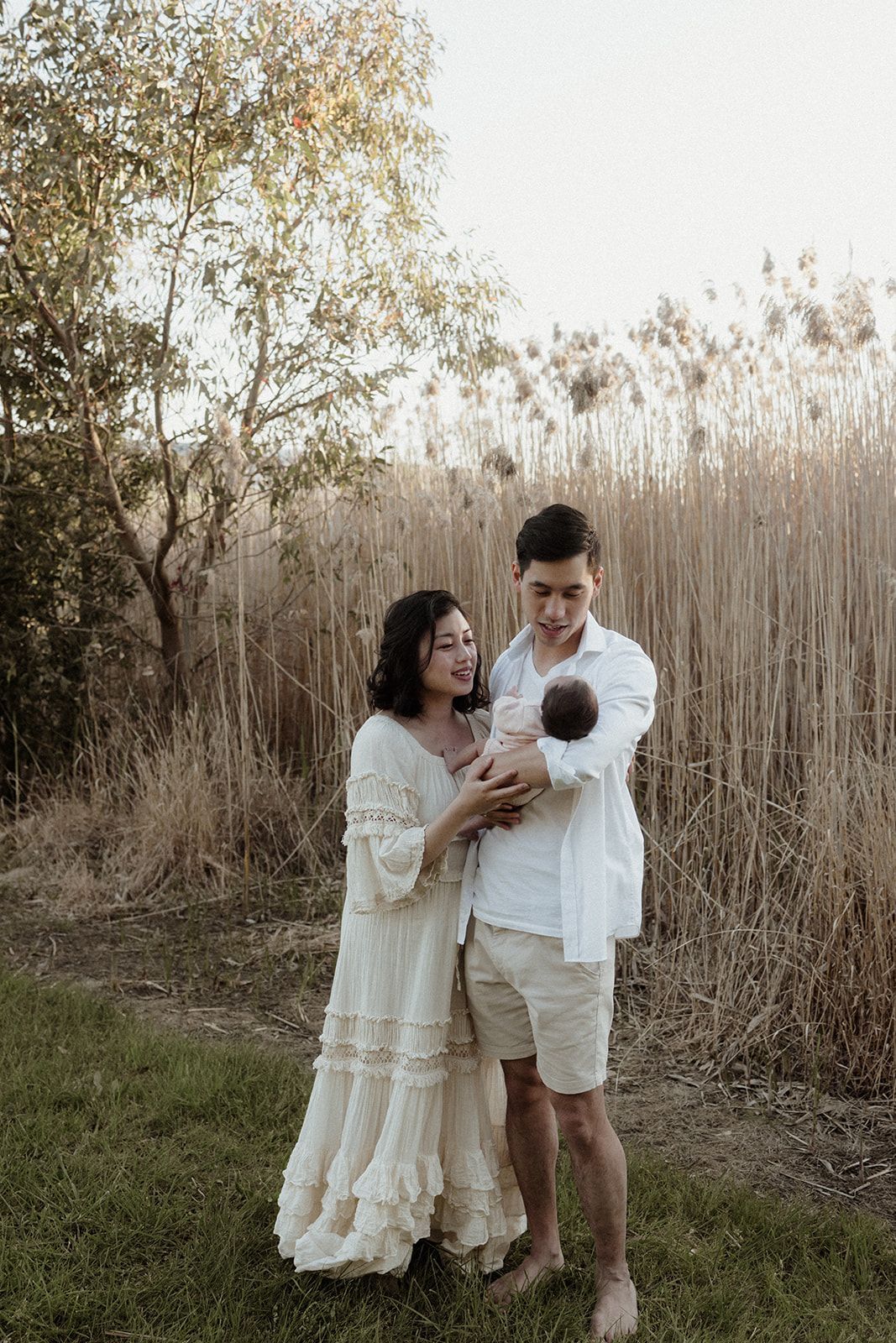 Couple in white outfits holding newborn baby outdoors near tall dry grass and trees during golden hour.