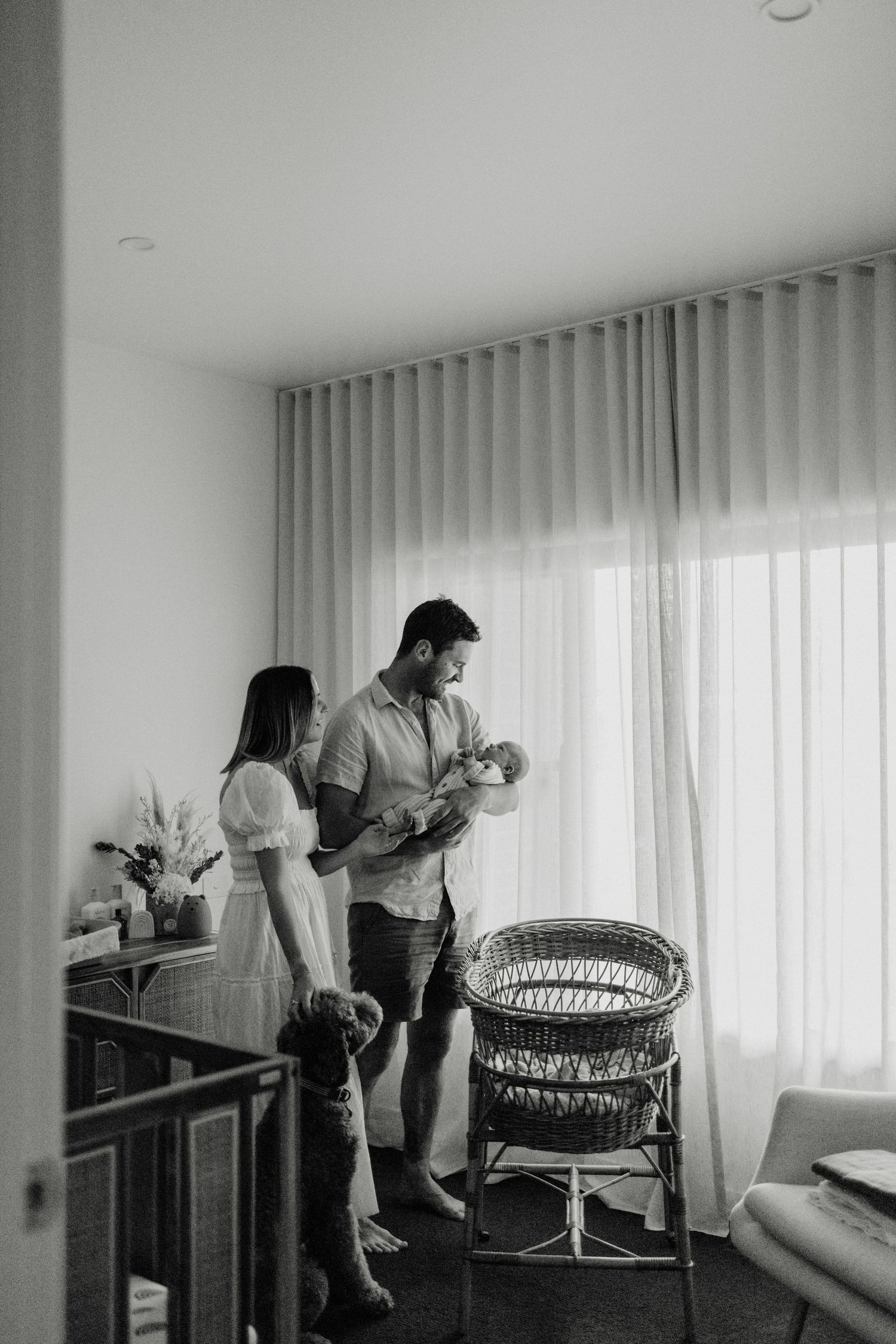 Black and white photo of parents holding their newborn baby in a bright nursery room with sheer curtains.
