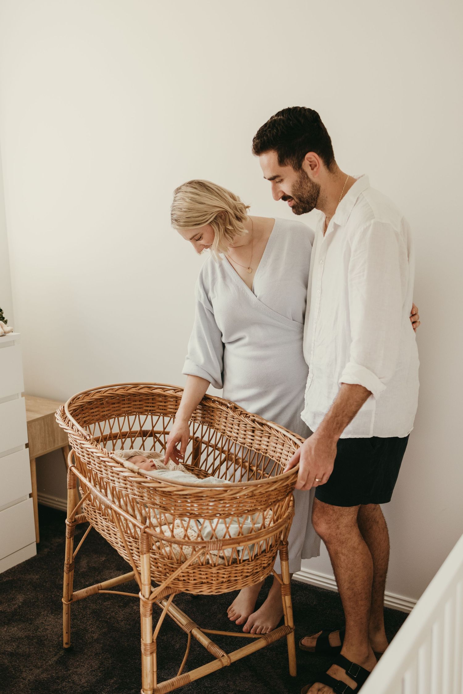 A couple in white clothing standing next to a vintage wicker bassinet in a minimalist nursery.