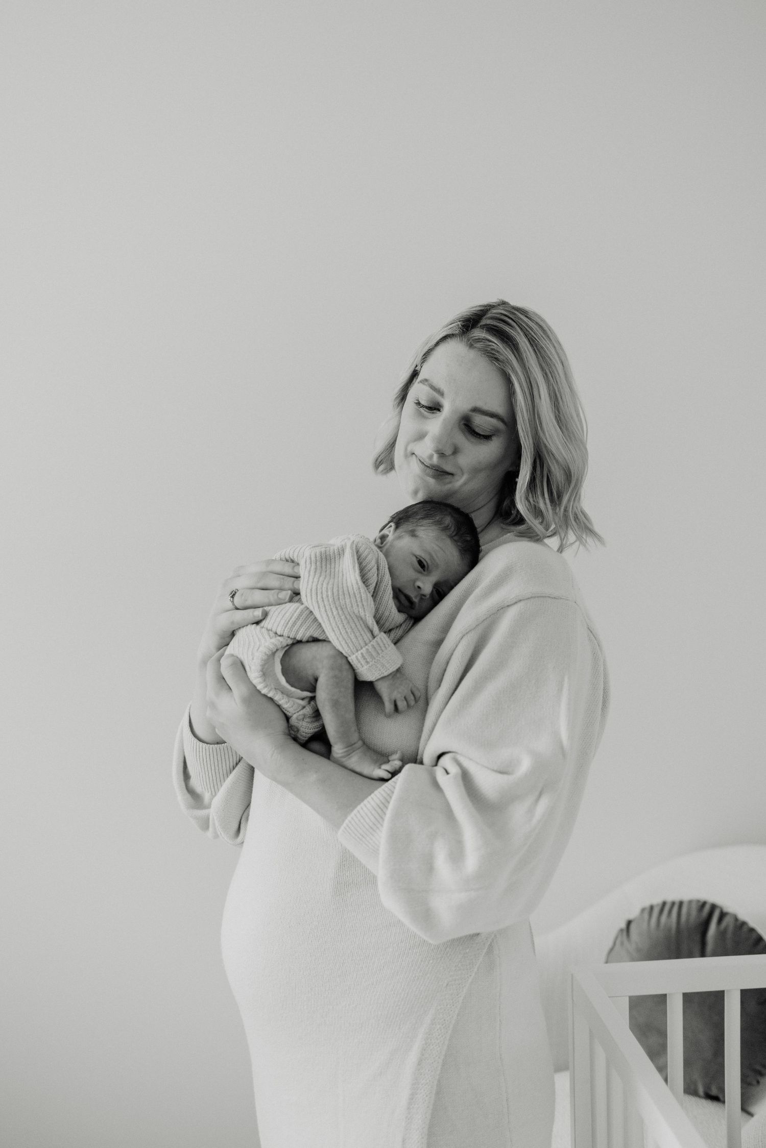 Black and white portrait showing a nurturing moment between parent and infant in a minimalist nursery setting.