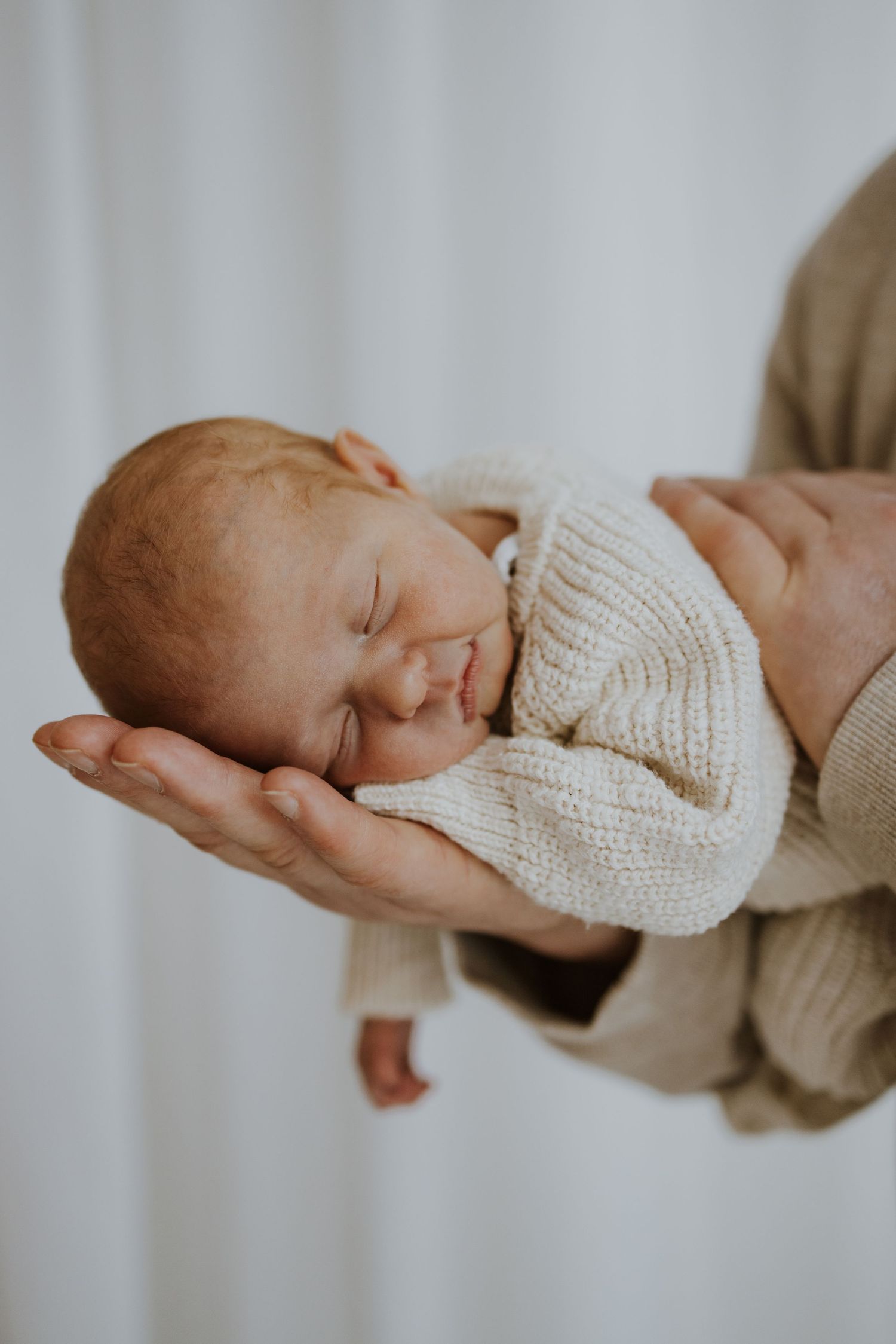 Newborn baby sleeping peacefully in parent's hands, wearing a cozy cream knit outfit against a white background.