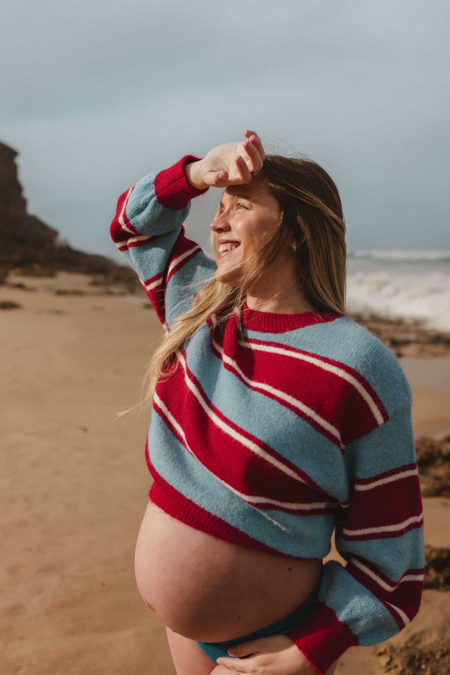 Smiling pregnant woman in striped sweater shielding eyes at beach, baby bump exposed, waves and rocky cliffs in background.