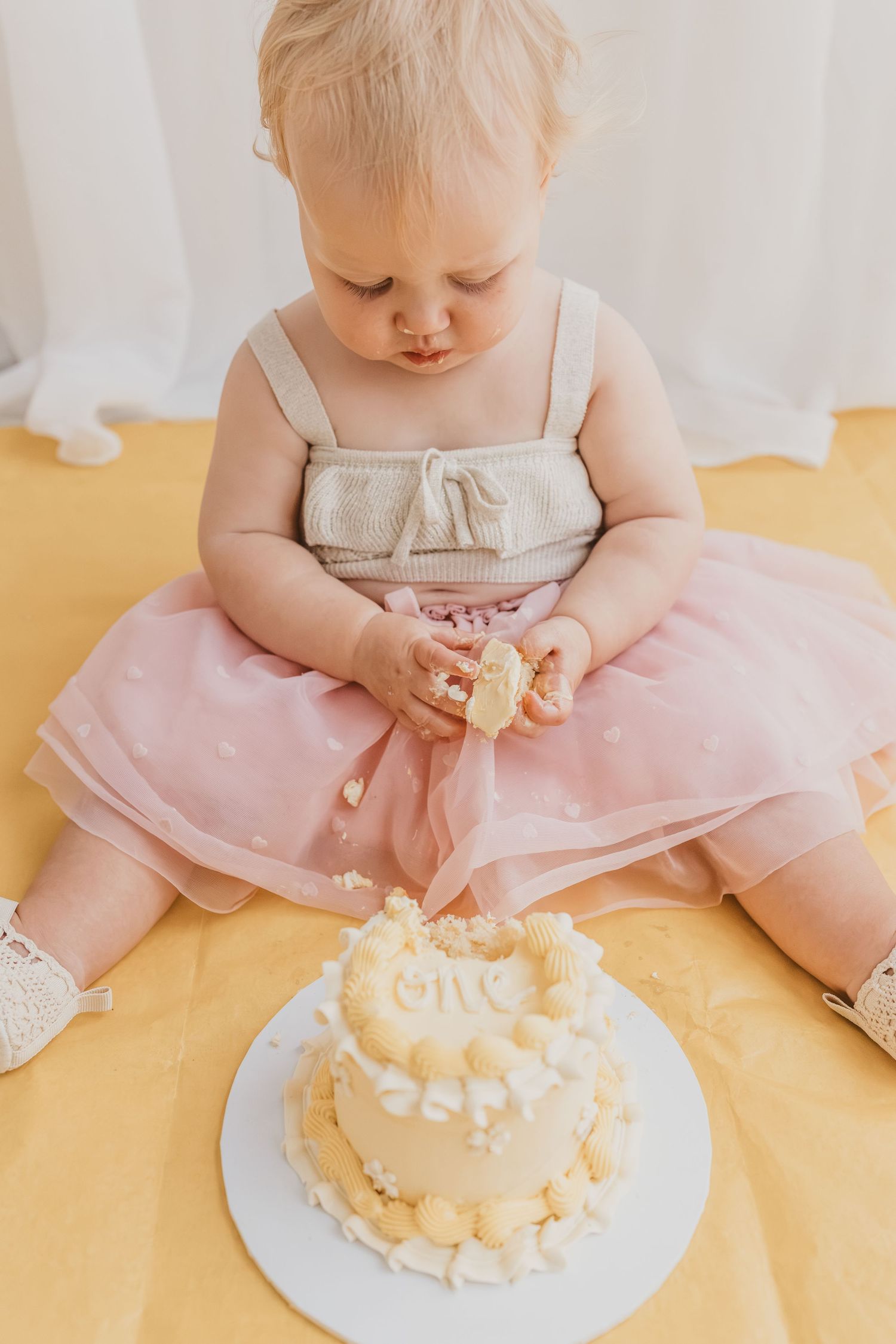 Baby girl in pink tutu dress smashing her first birthday cake, sitting on yellow background.