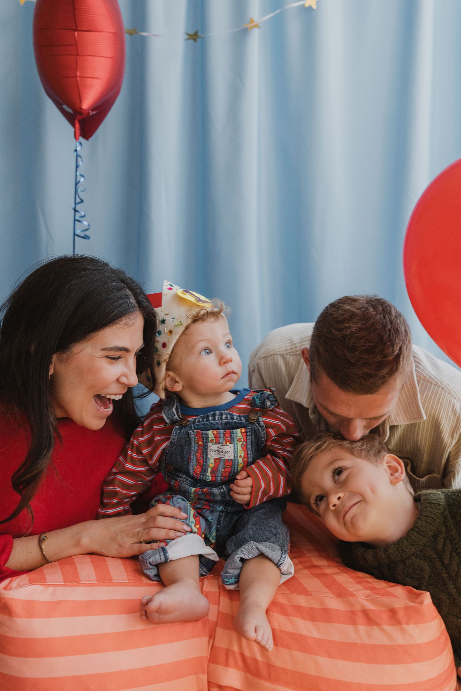 Family of four celebrating a birthday with red balloons, parents smiling at baby wearing party hat on colorful cushions.