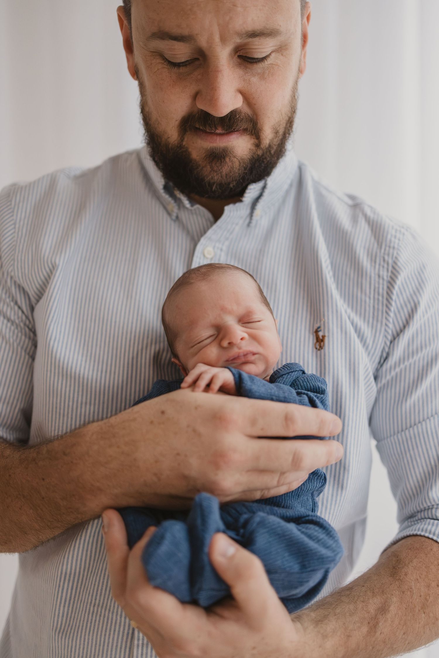 A bearded father in a striped shirt tenderly holds his sleeping newborn baby wrapped in a blue swaddle.