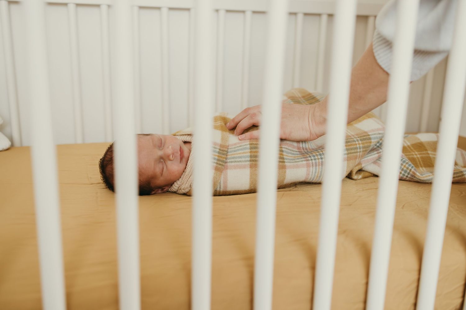 Sleeping baby wrapped in plaid blanket viewed through white crib railings.