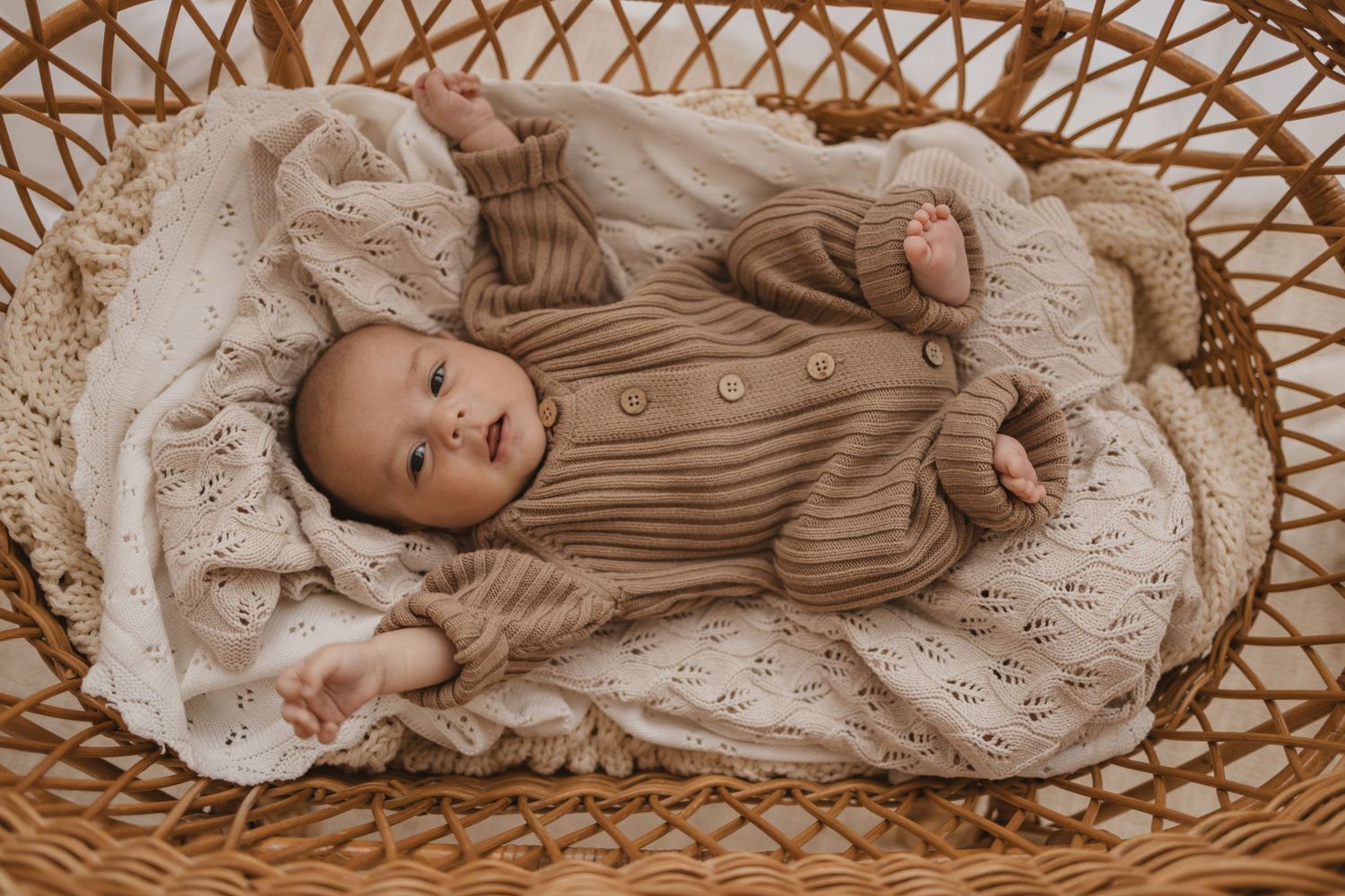 Baby in tan knit romper lying in wicker bassinet with cream lace blanket, smiling softly.