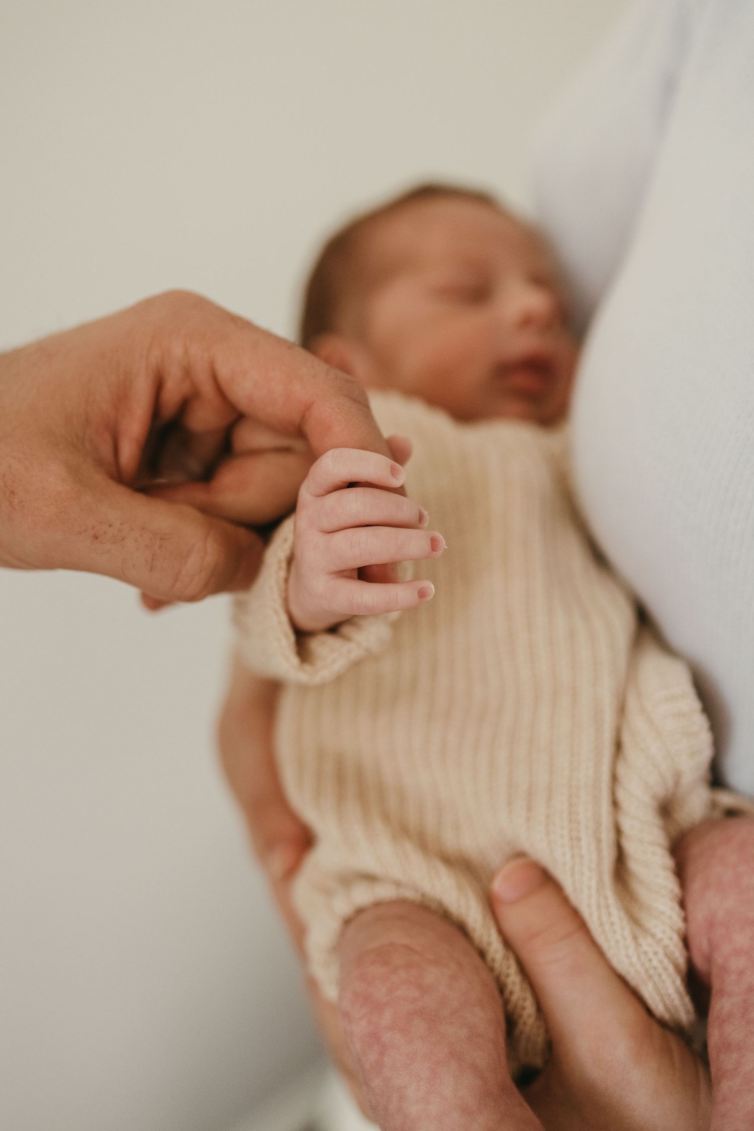Newborn baby in cream knit outfit holds parent's finger in intimate bonding moment.