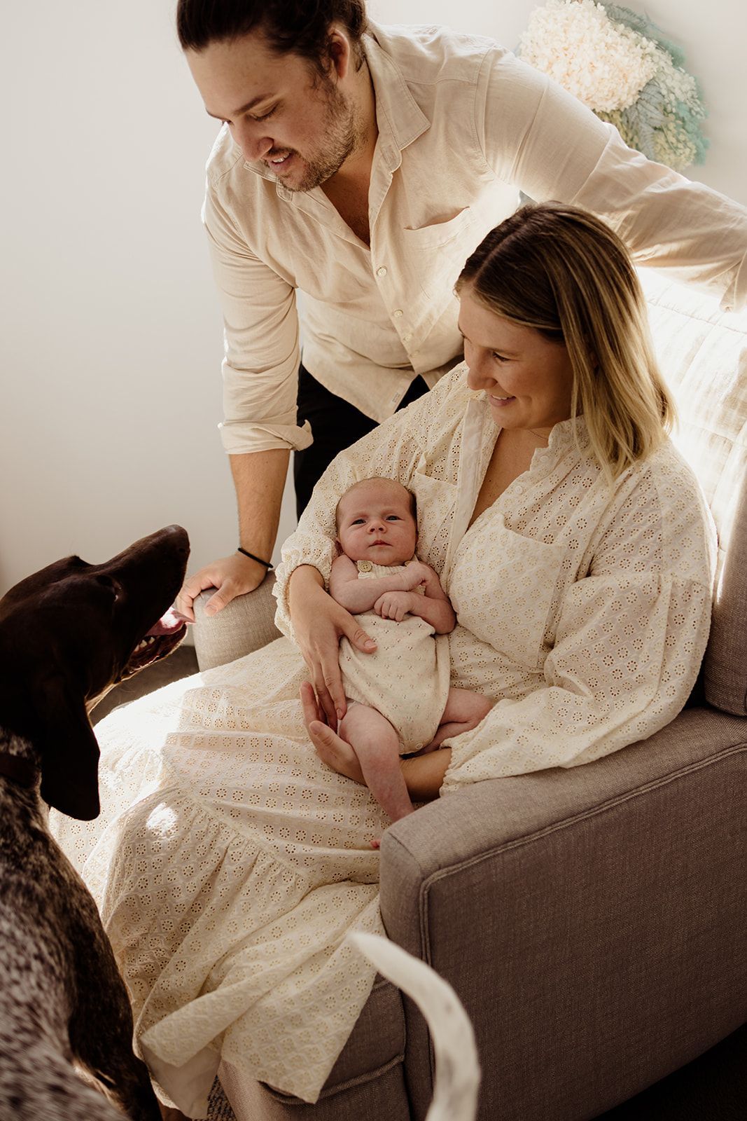 A couple in white outfits smile at their newborn baby while a dog sniffs the infant in a cozy room.