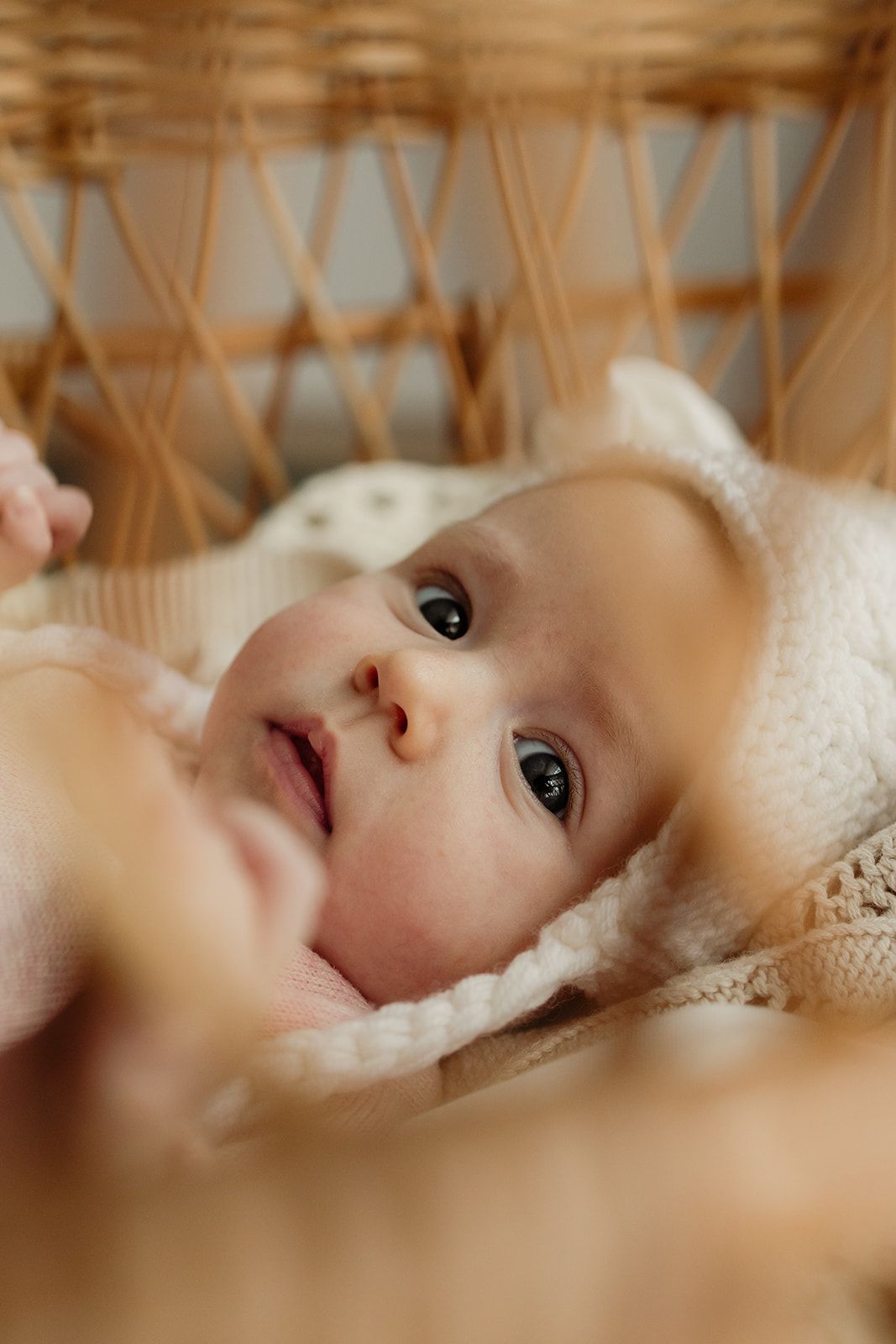 Newborn baby lying in a wicker bassinet wearing a cream knit bonnet, looking up with wide dark eyes.