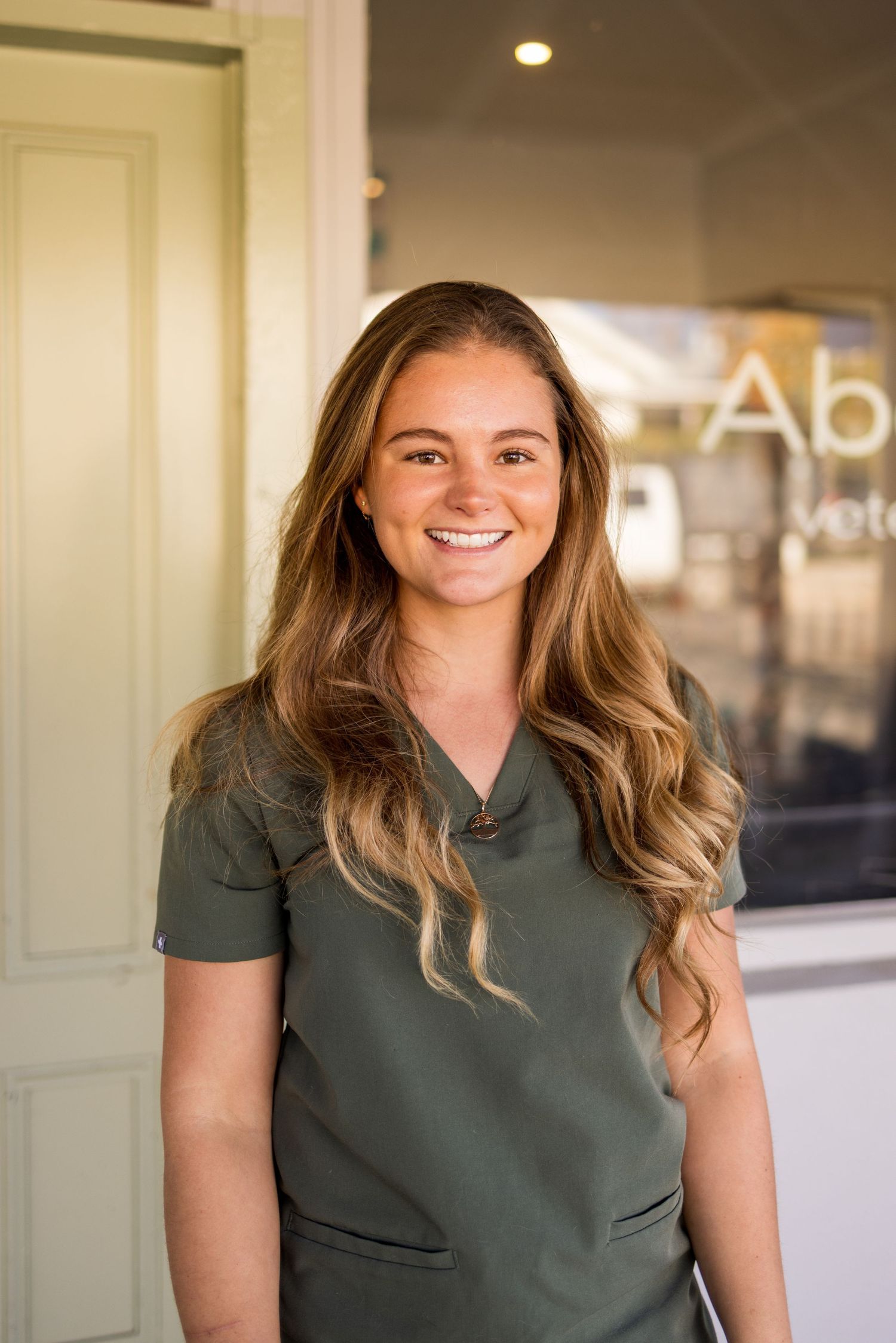 Smiling young woman in olive green scrubs standing in front of a veterinary clinic entrance.