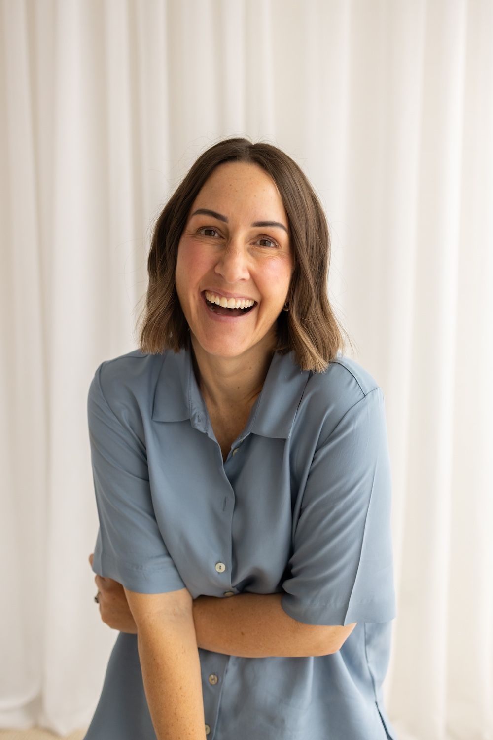 A smiling woman with brown hair in a blue button-up shirt poses with arms crossed against a white curtain backdrop.