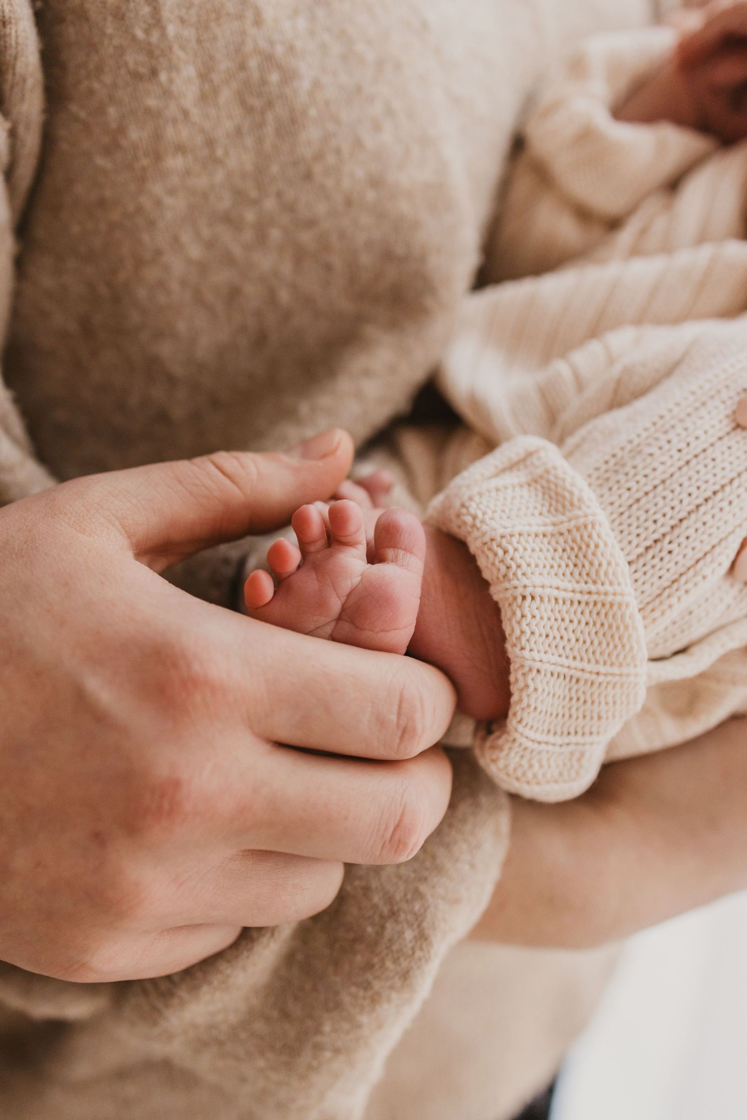 Close-up of a parent gently holding a newborn baby's tiny feet in their hands, wearing a cozy beige knit sweater.