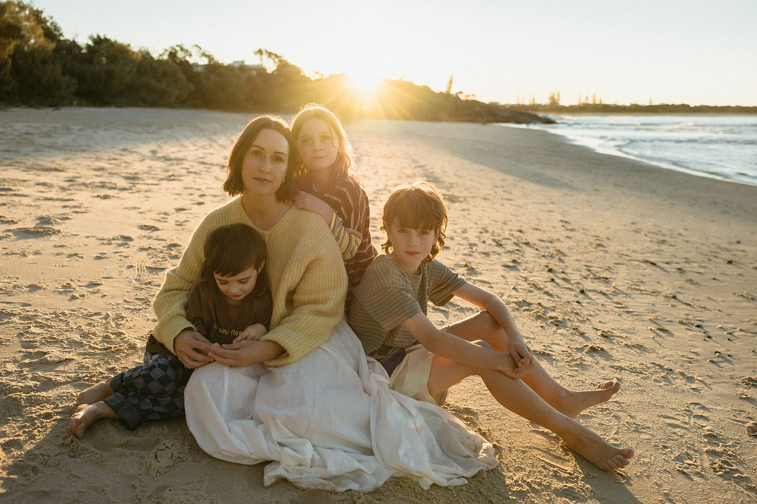 Family of four sitting together on a sandy beach at golden sunset, two adults and two children smiling warmly.