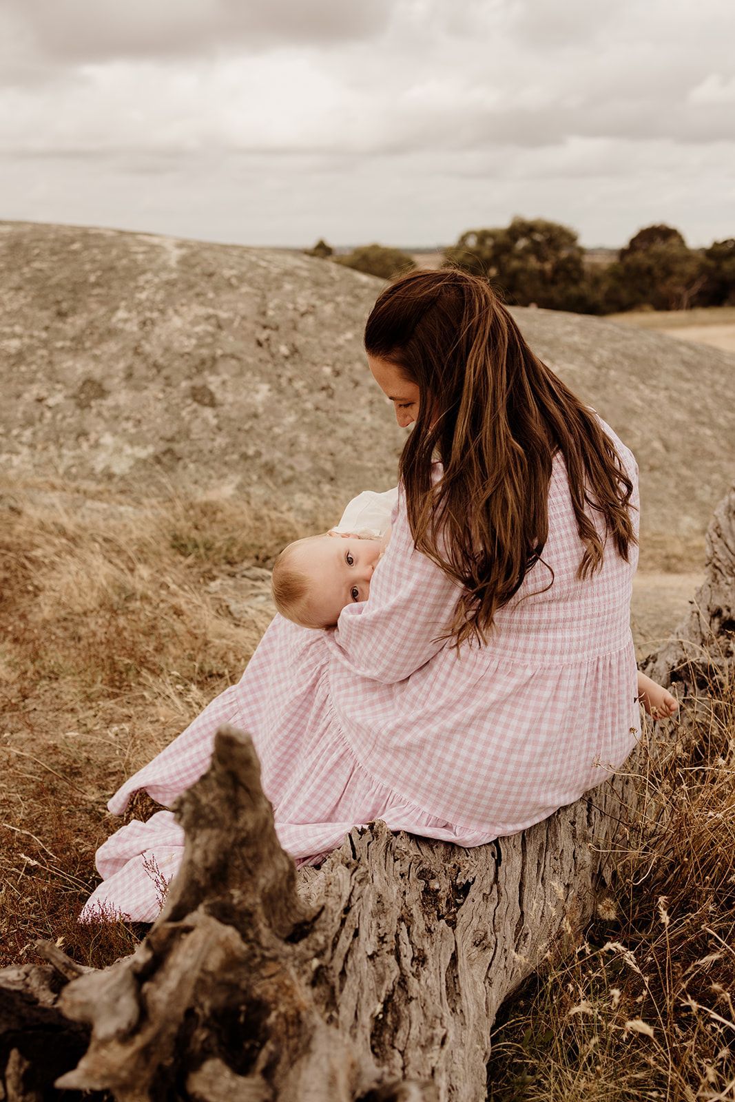 A mother in a pink gingham dress sits on a log breastfeeding her baby in a scenic outdoor setting.