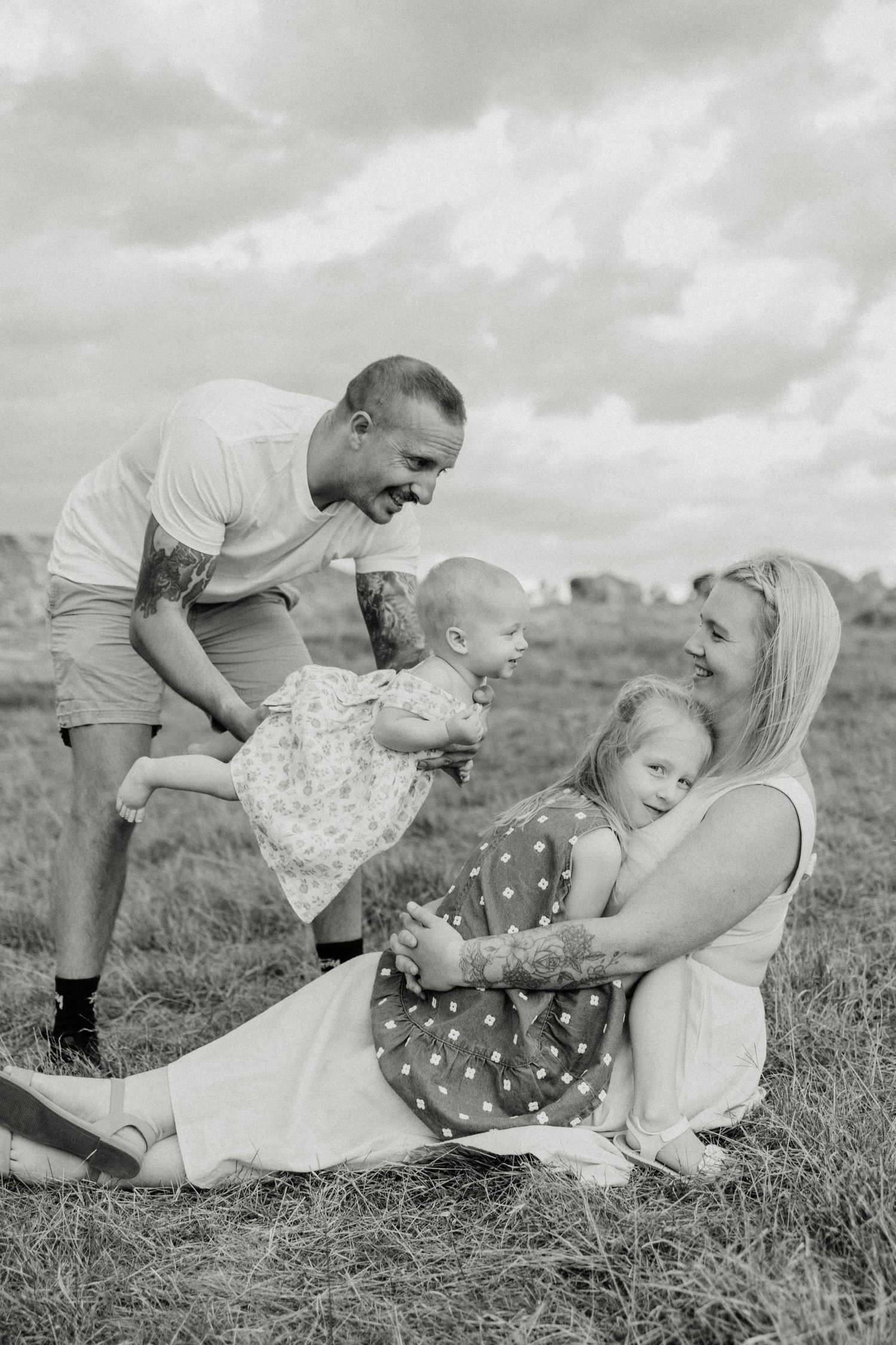 Black and white family photo of parents playing with two young daughters in a grassy field under a cloudy sky.