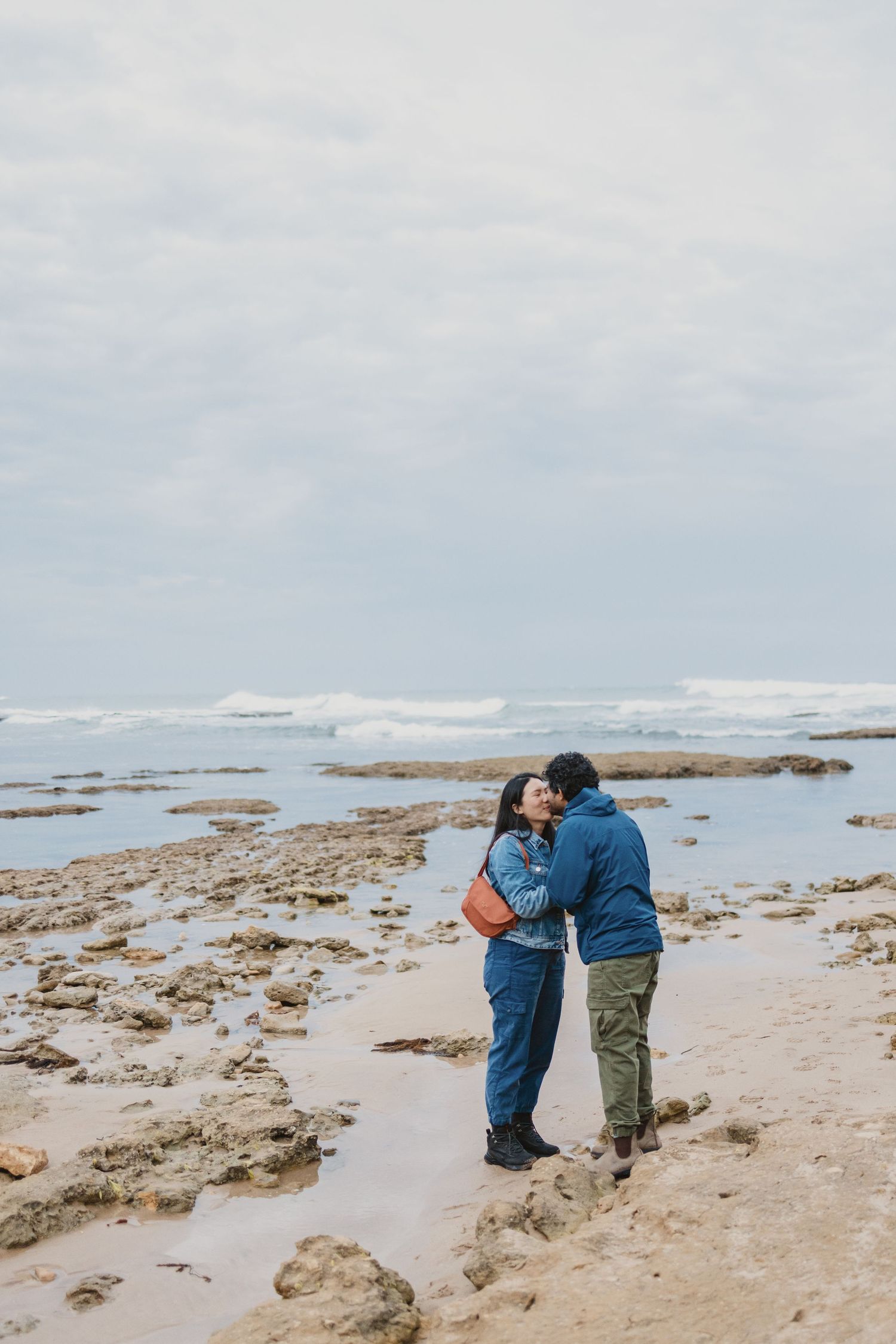 Couple sharing a romantic kiss on a rocky beach with ocean waves in the background on a cloudy day.