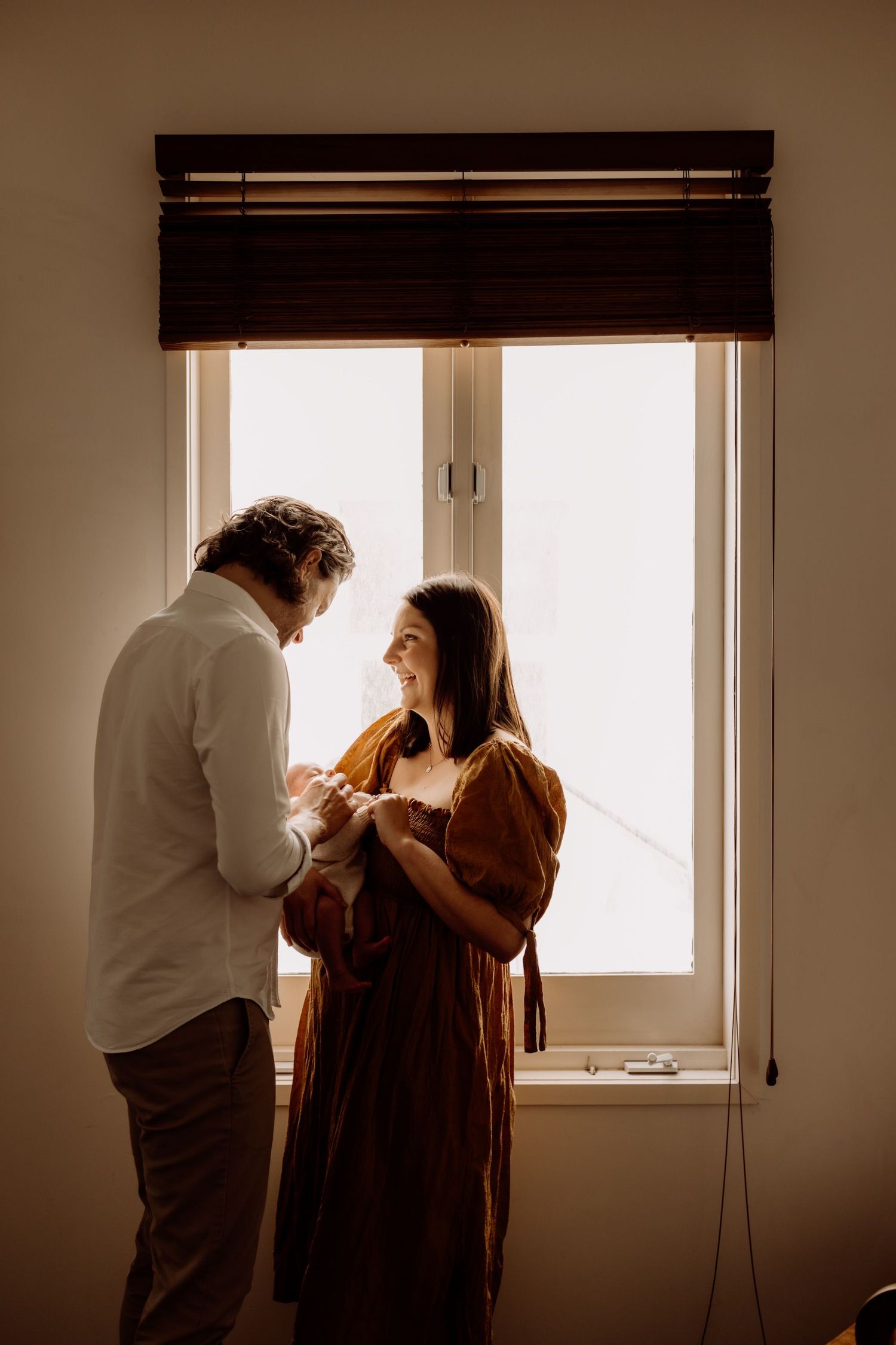 A couple stands by a sunlit window, the woman in a rust dress holding a newborn baby while smiling at her partner.