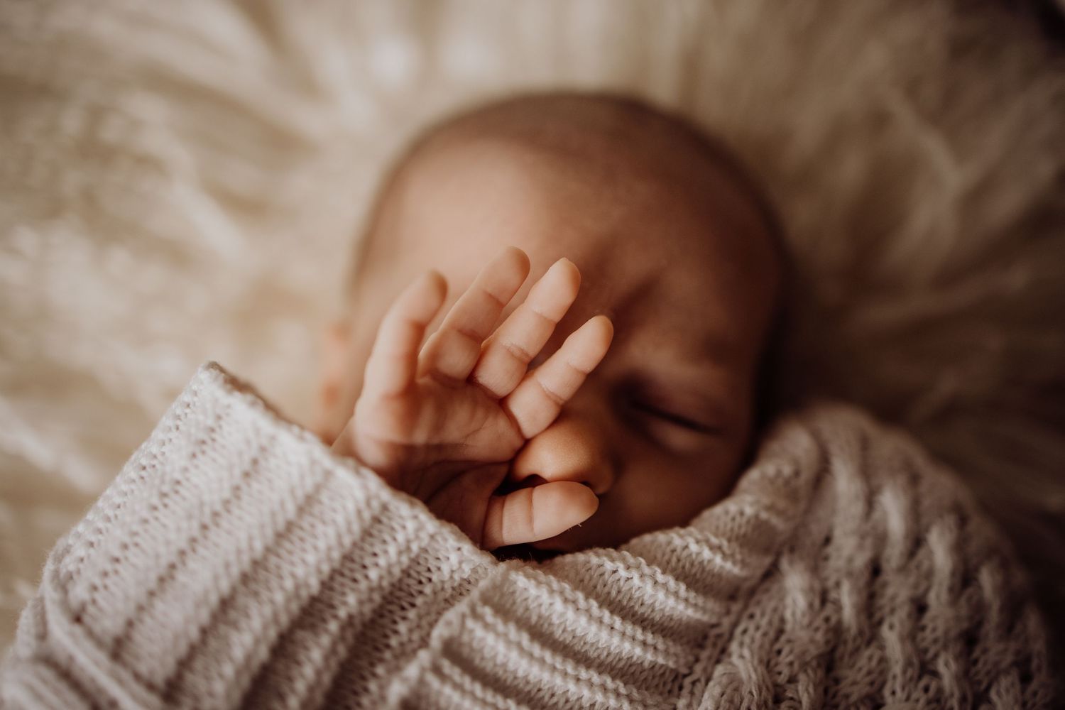 Sleeping newborn baby wrapped in a cozy knit blanket with tiny hand resting gently on face.