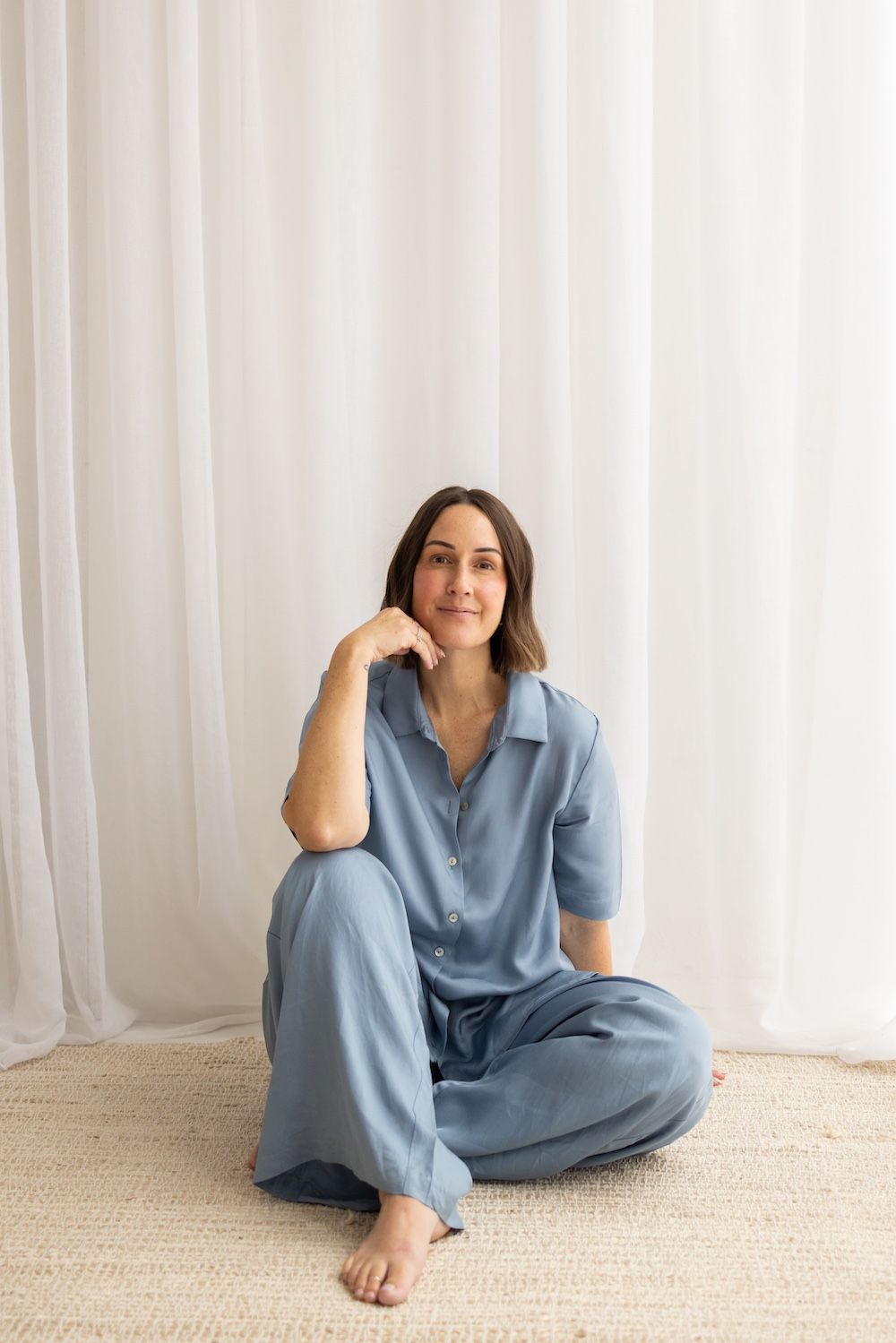 Woman sitting cross-legged on a rug wearing a blue satin short-sleeve button-up shirt and matching wide-leg pants.