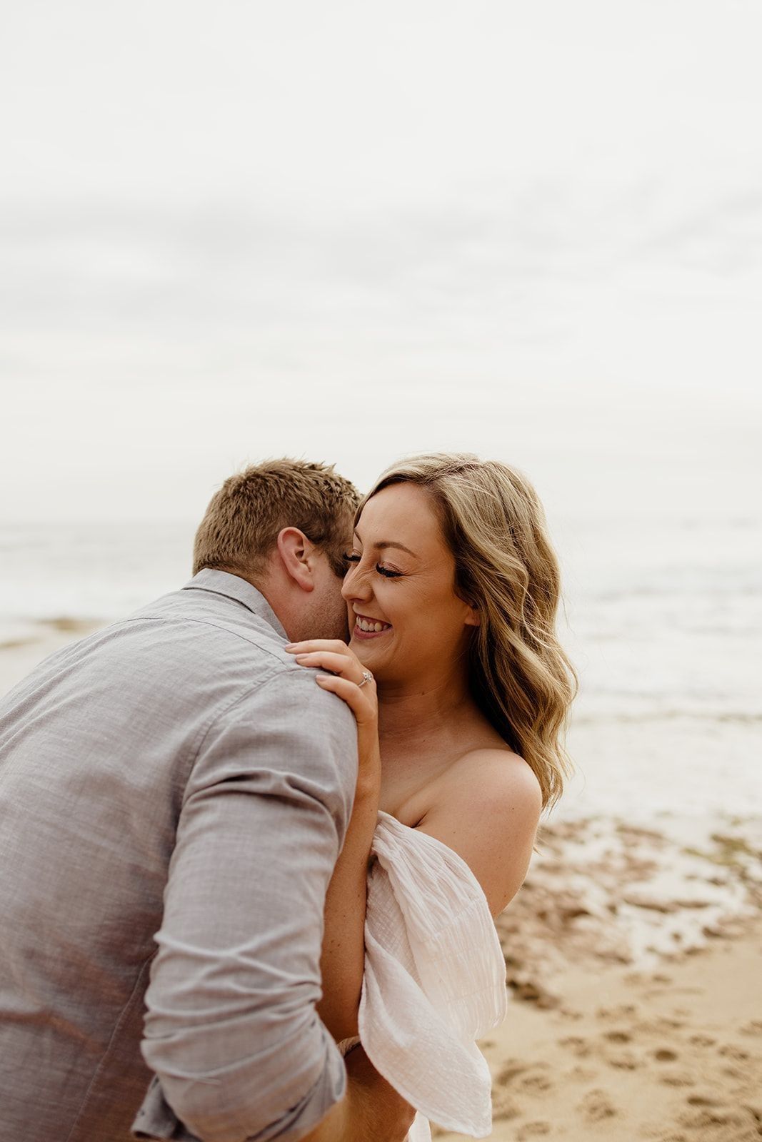 A couple shares a romantic beach moment, the groom whispering to his smiling bride in a white dress by the ocean.