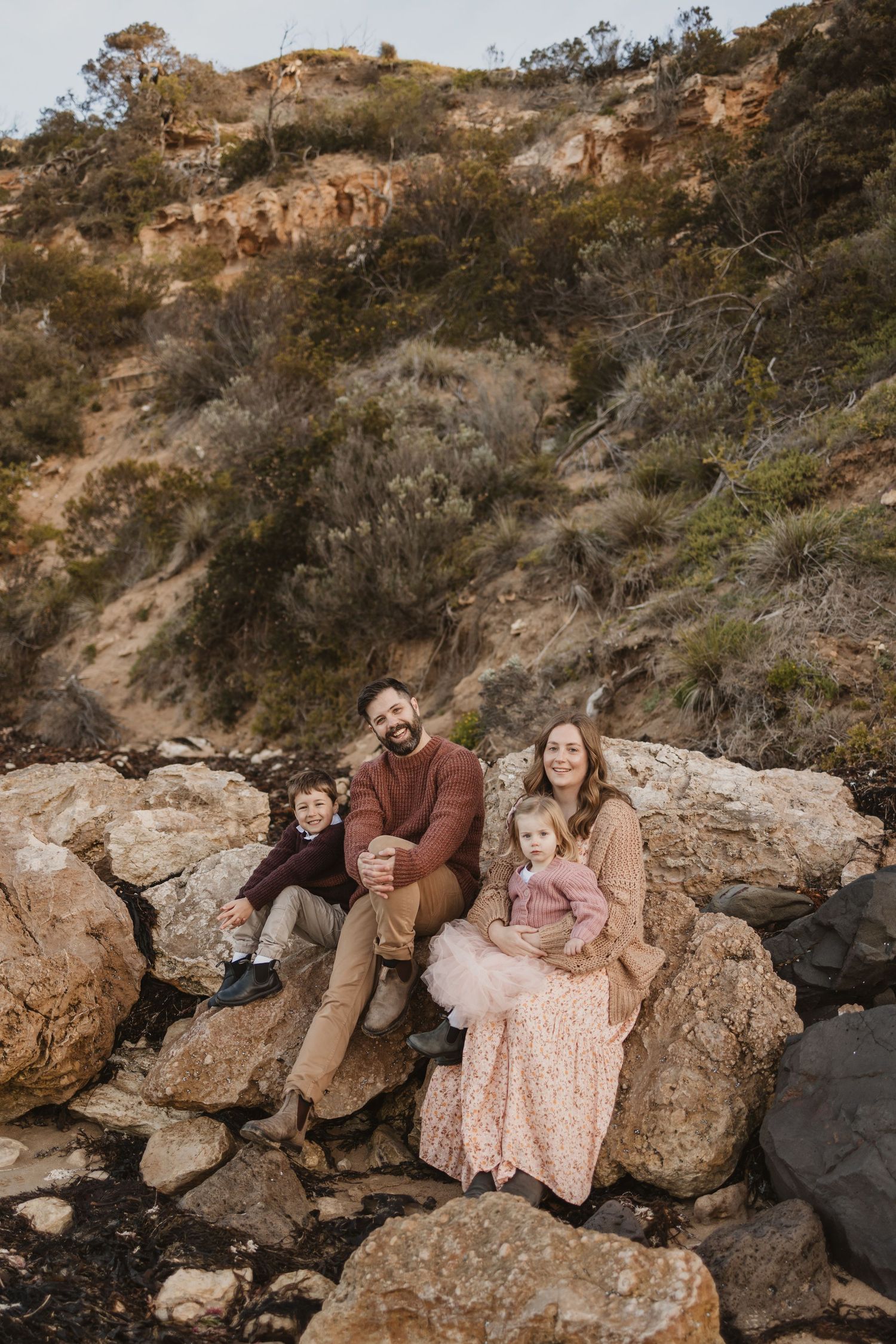 Family of four sitting on rocky terrain with a hillside backdrop, smiling warmly in earthy tones.