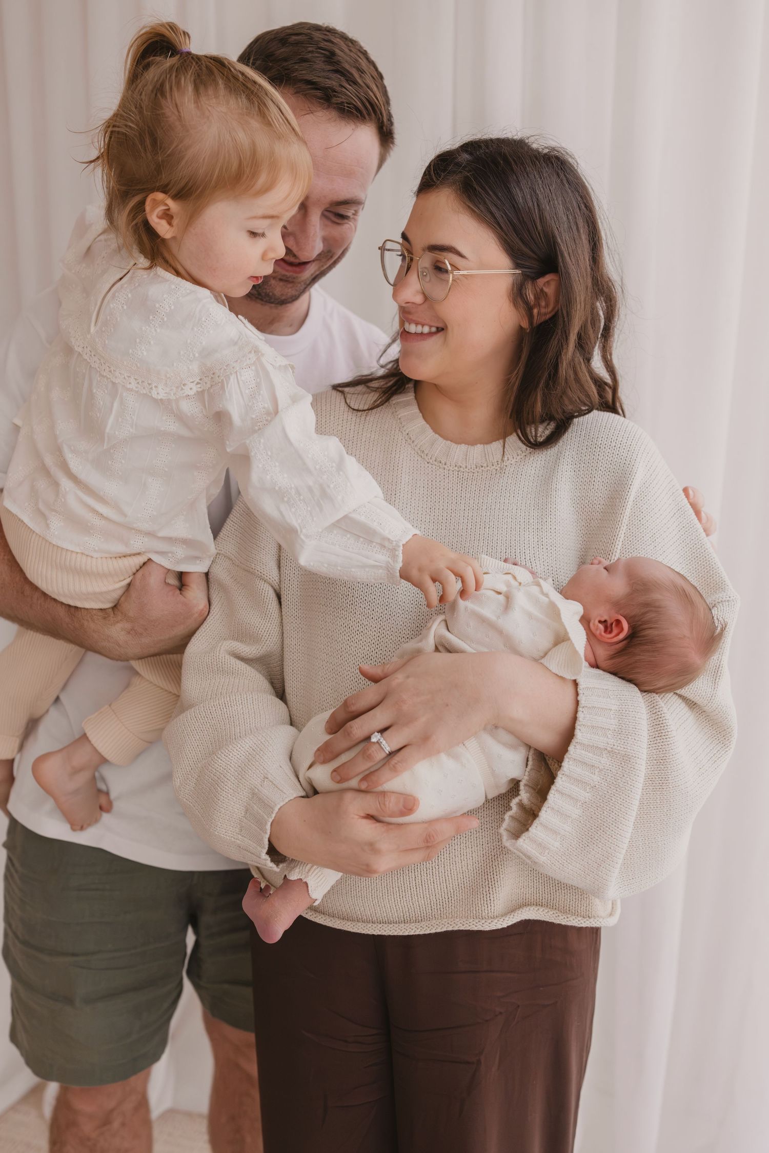 Happy family of four with parents holding toddler and newborn baby, wearing neutral tones in bright studio setting.