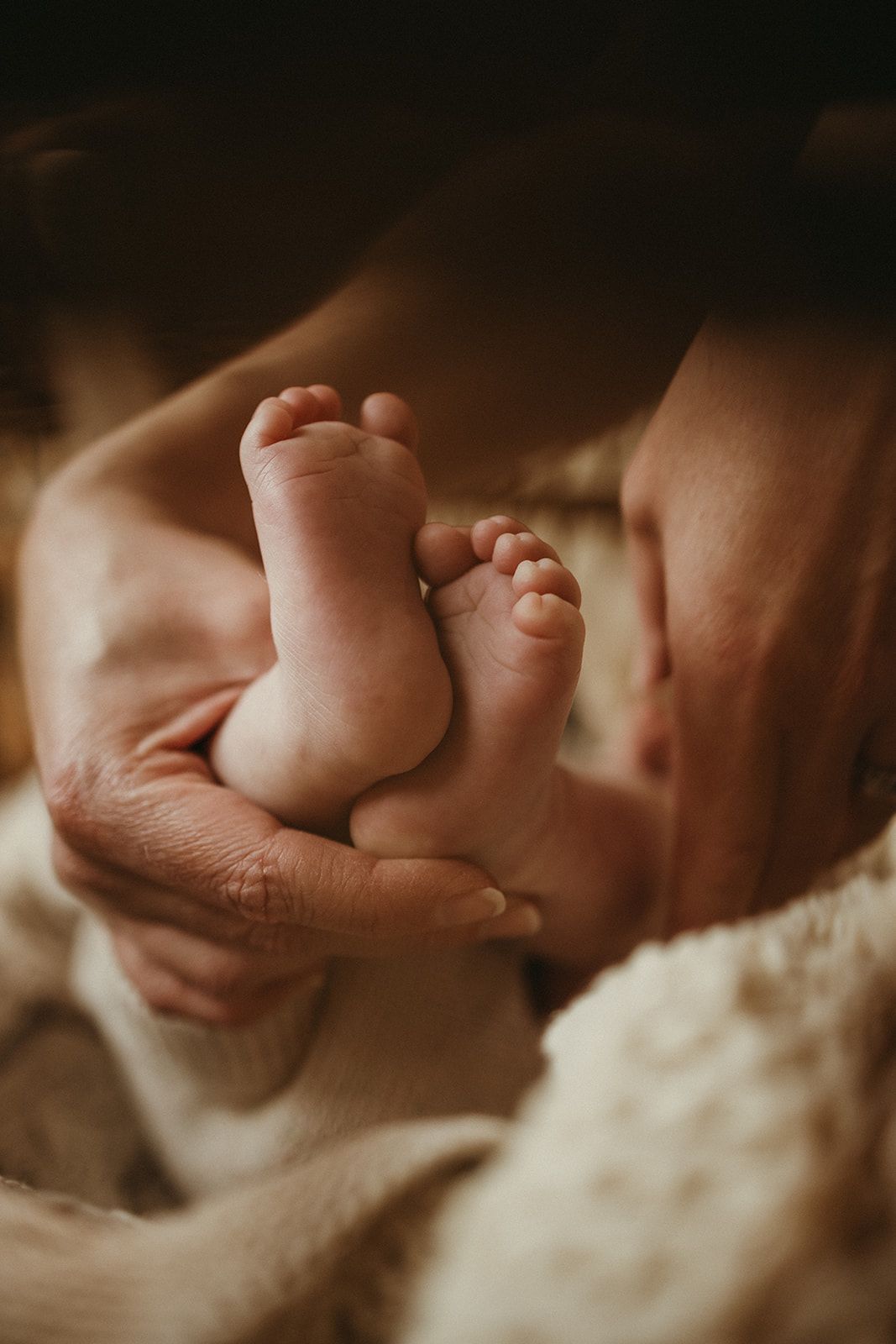 Adult hands gently cradling tiny newborn baby feet wrapped in a soft cream blanket in warm light.