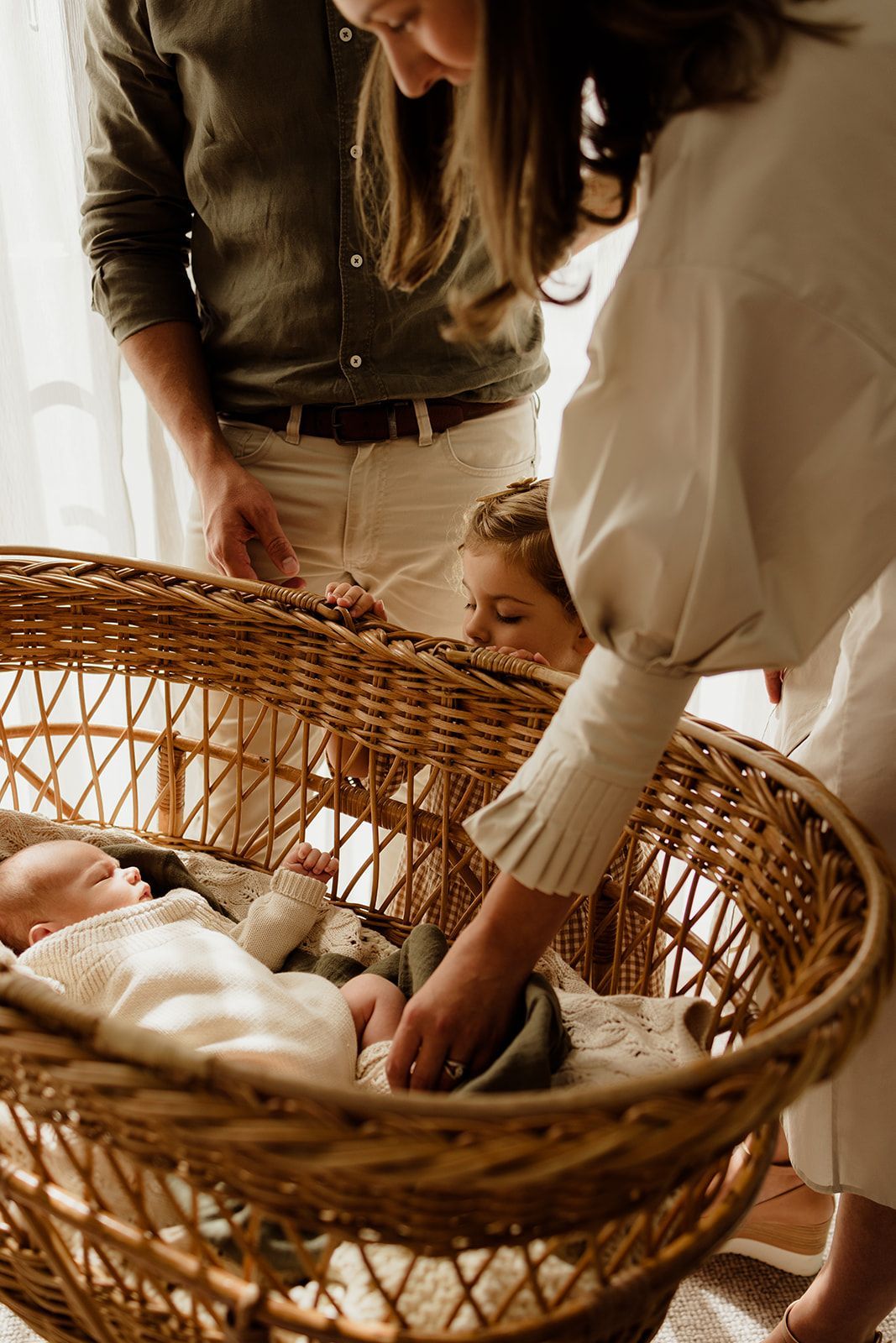 Family gathered around a wicker bassinet with newborn baby, parents and toddler sharing a tender moment.